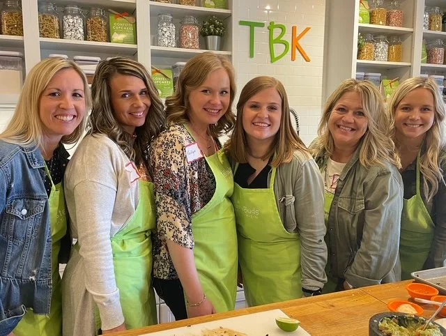 Six women standing together in a kitchen with shelves of spices behind them, wearing green aprons and smiling for the camera.
