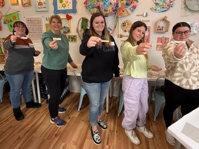 Five women standing in a room decorated with colorful artwork, each holding a small piece of paper or craft item, smiling and pointing towards the camera.
