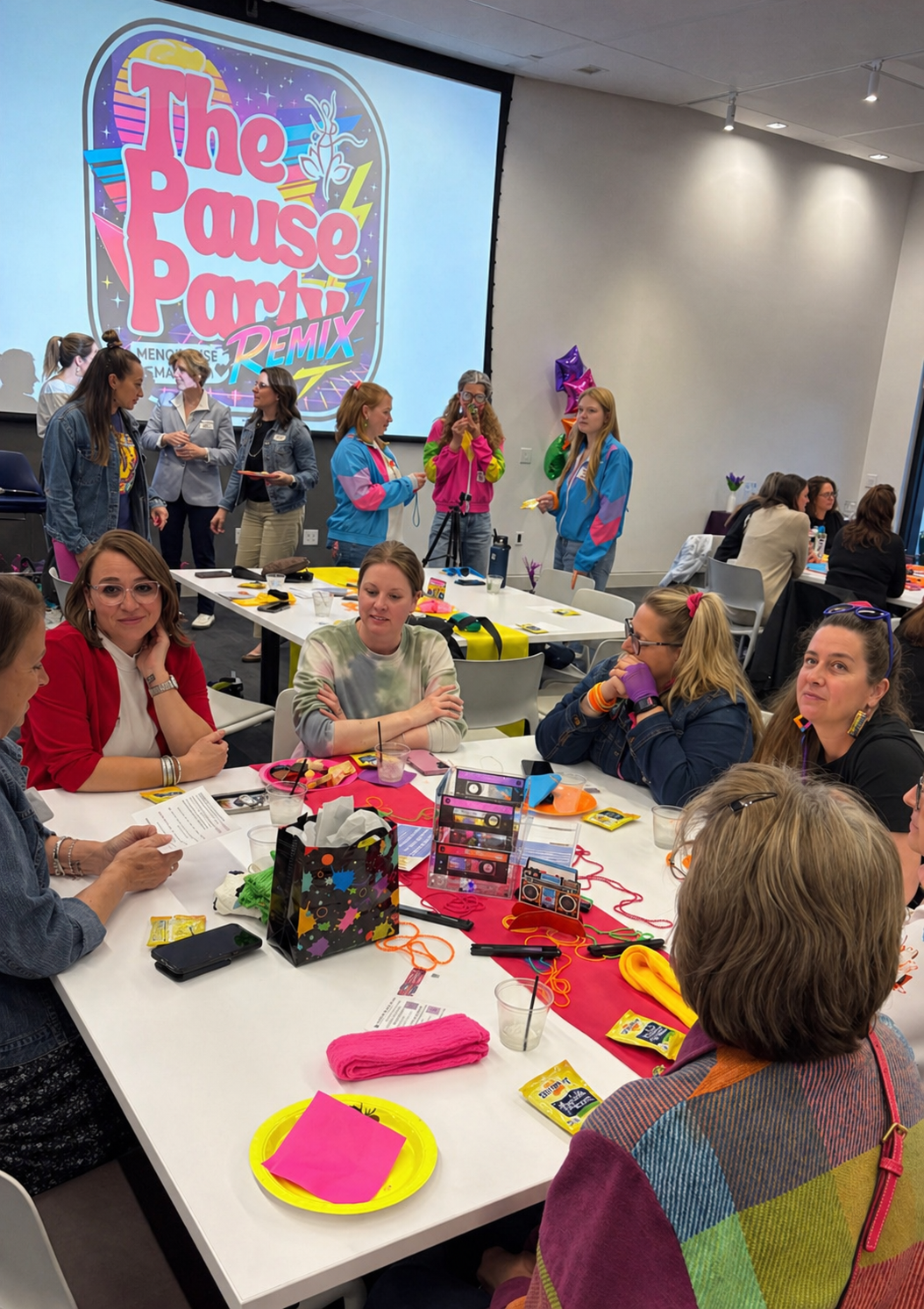 A group of women sitting around a table at a party, with some women standing near a large screen that displays a colorful "The Pale Party Remix" logo. The table is decorated with party supplies, including a gift bag, pink napkins, and snacks.