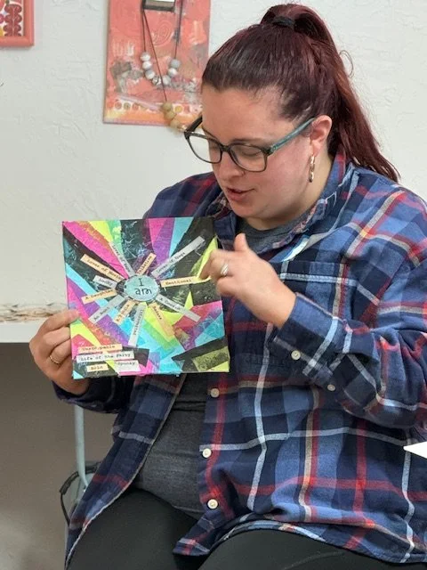 Woman with glasses and a plaid shirt looking at a colorful scrapbook or card with a rainbow design and various cut-out words and images.