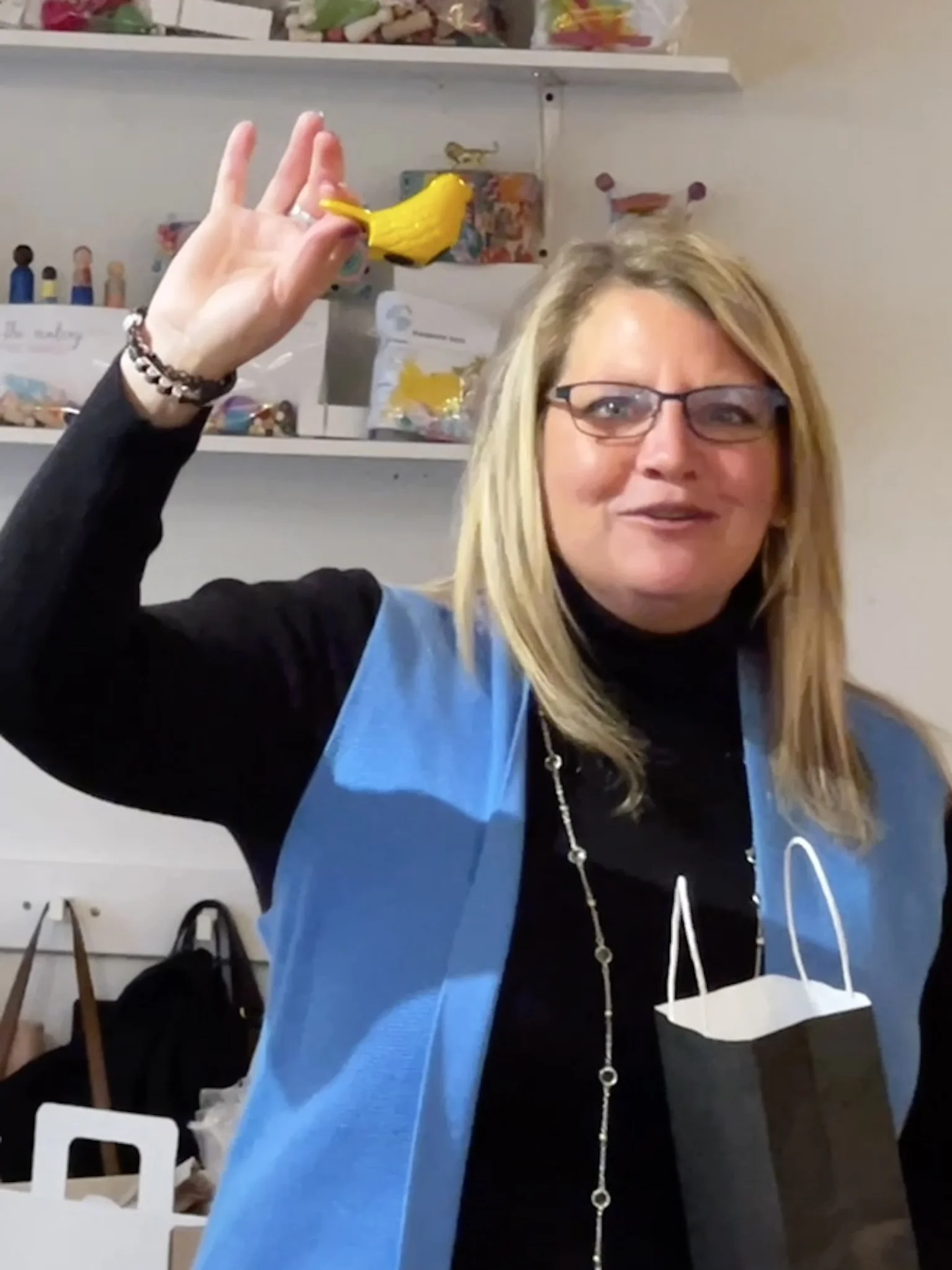 A woman with blonde hair, glasses, and a black top wearing a blue apron, holding a small yellow bird-shaped object in her right hand. She is smiling, and there are shelves with colorful items in the background, along with a white table and shopping bags.