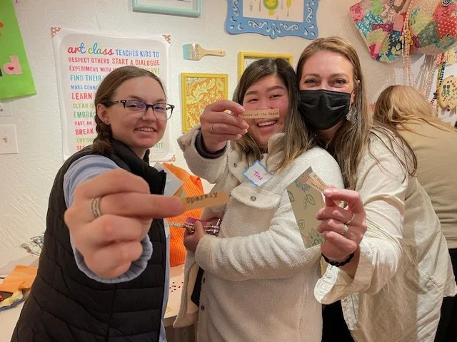 Three women smiling at a classroom craft event, holding small art projects, with colorful wall decorations and posters in the background.