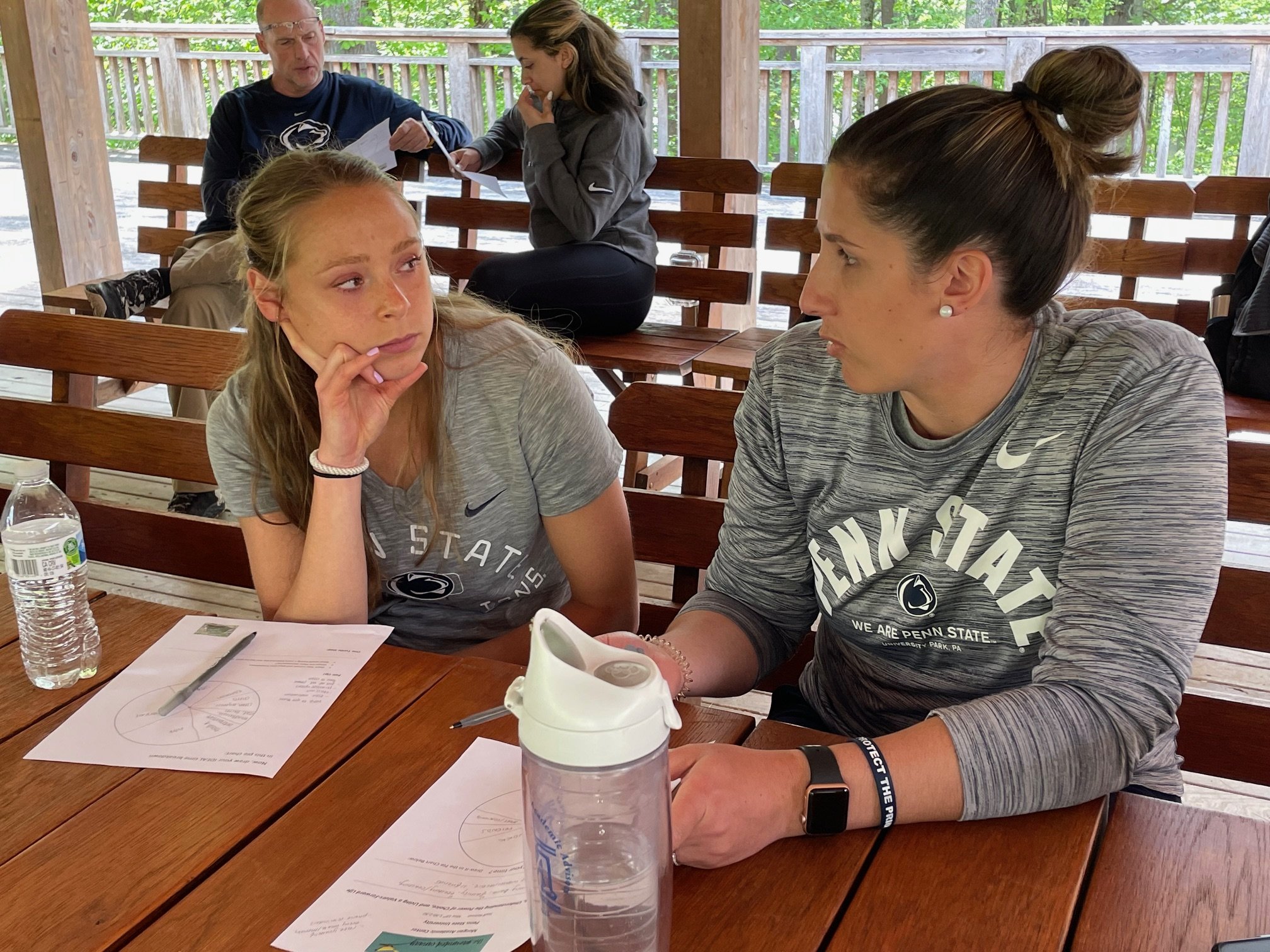 Two young women and an older man seated at a wooden table outdoors, engaged in conversation and paperwork, with two women in athletic attire and notebooks in the background under a wooden shelter with green trees visible behind.