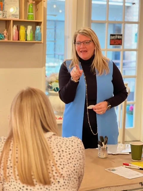 A woman with blonde hair and glasses, wearing a black top and light blue vest, standing in front of a window, giving a presentation or speech to a seated woman with long blonde hair.