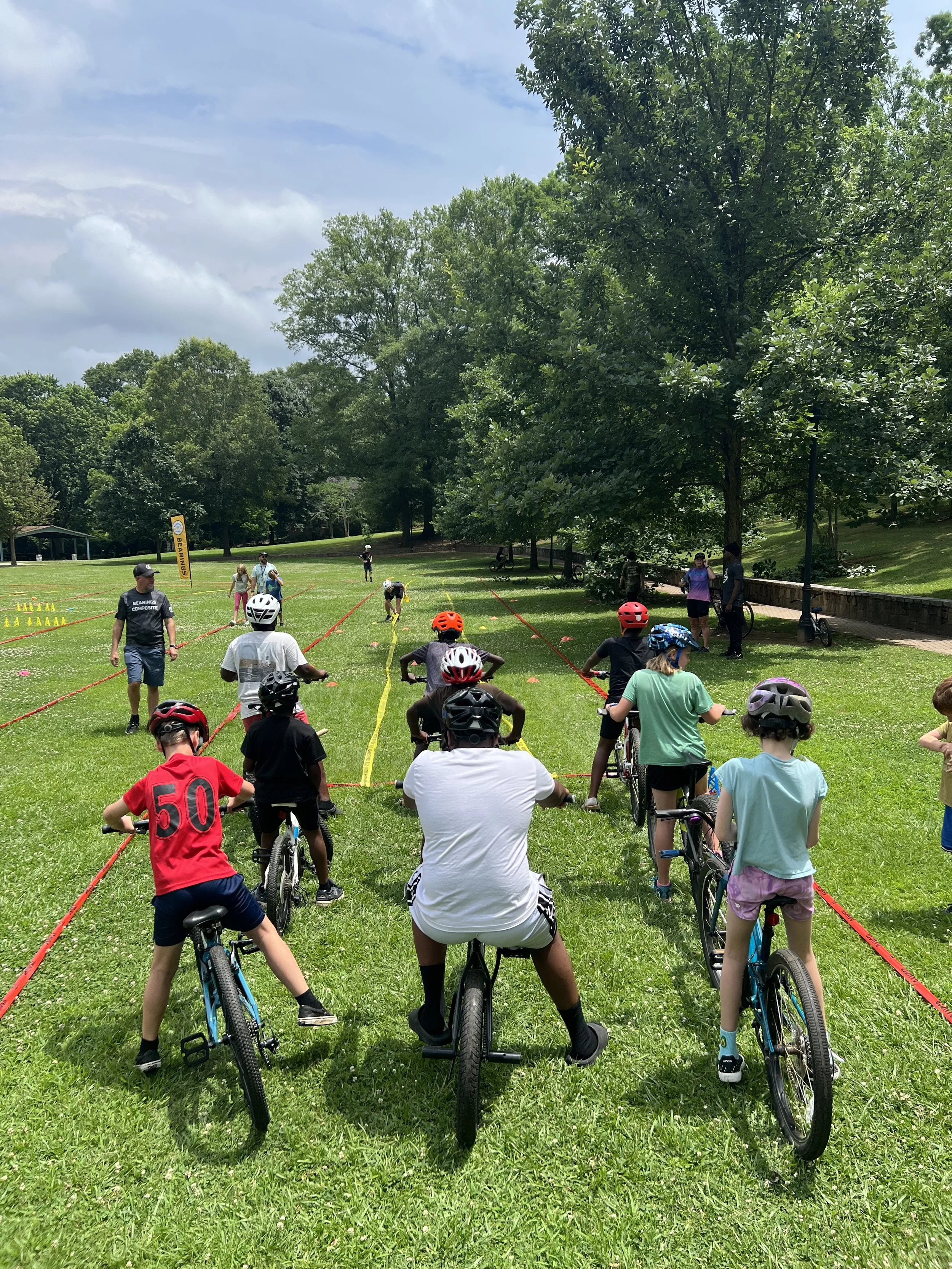 Group of children and adults with bikes participating in a bike riding class or event on a grassy field in a park, with trees and cloudy sky in the background.