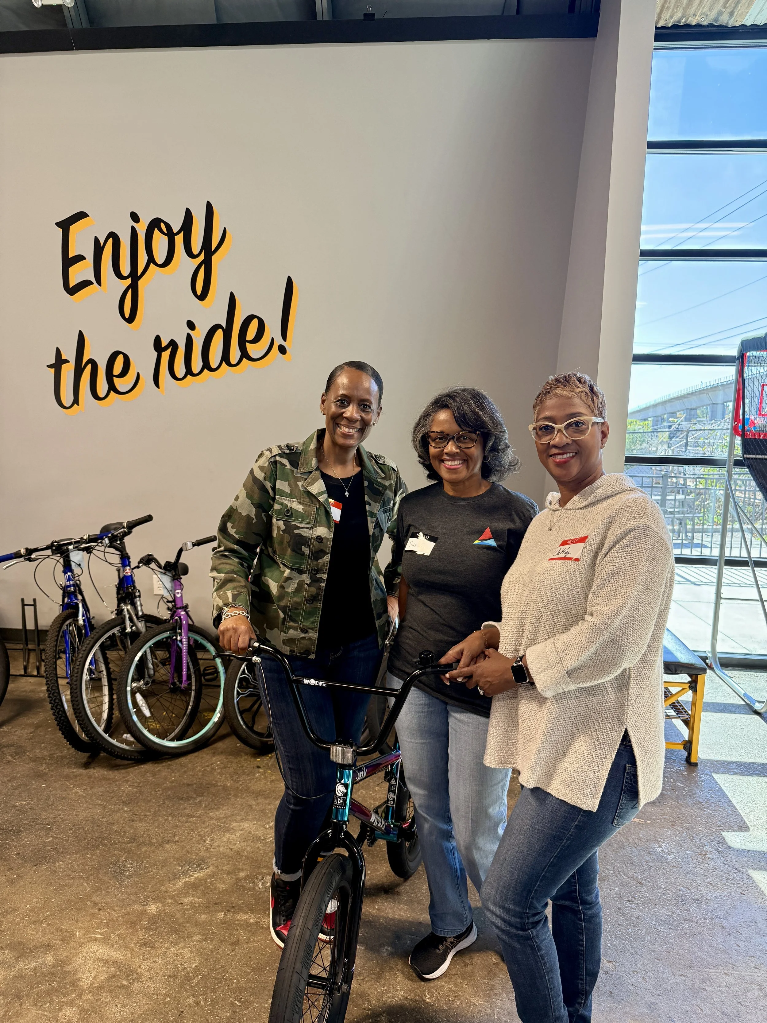 Three women standing indoors with bicycles, smiling, in front of a wall with a sign that says 'Enjoy the ride!'. One woman is holding a black bicycle, while others stand beside her.
