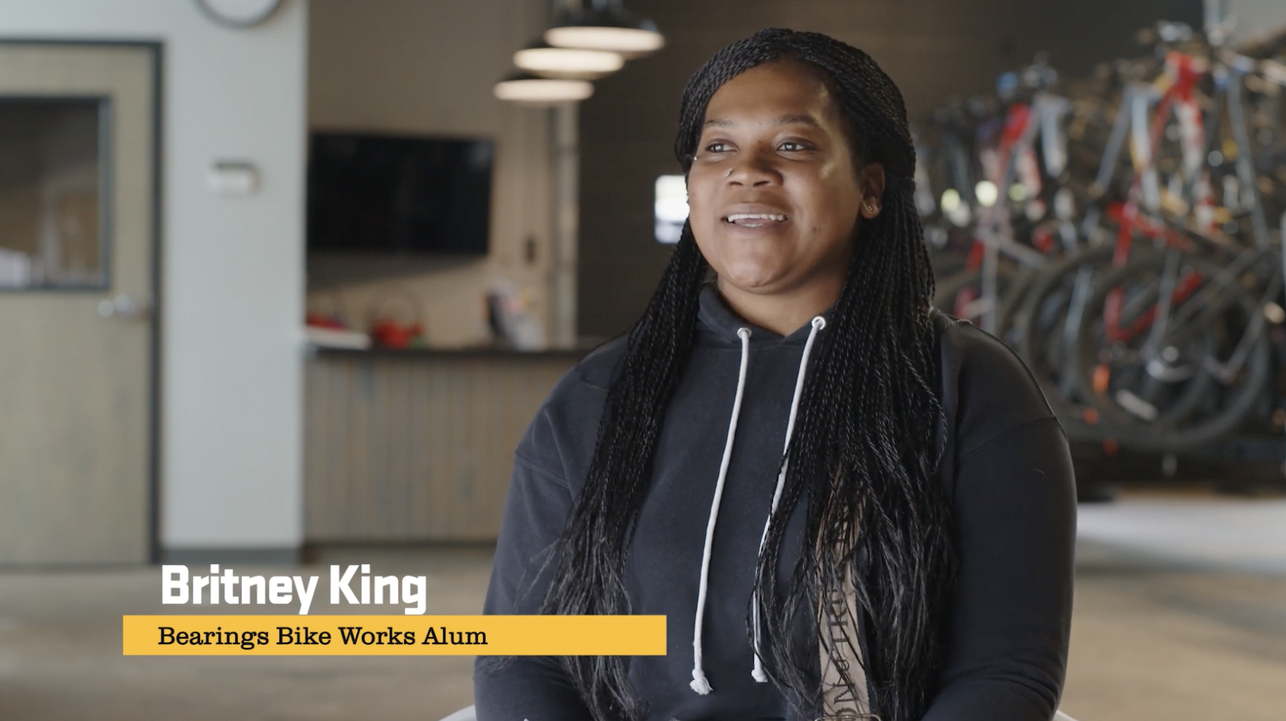 A woman with long braided hair and a black hoodie sitting in a bike shop or workshop. Bicycles are visible in the background on racks.