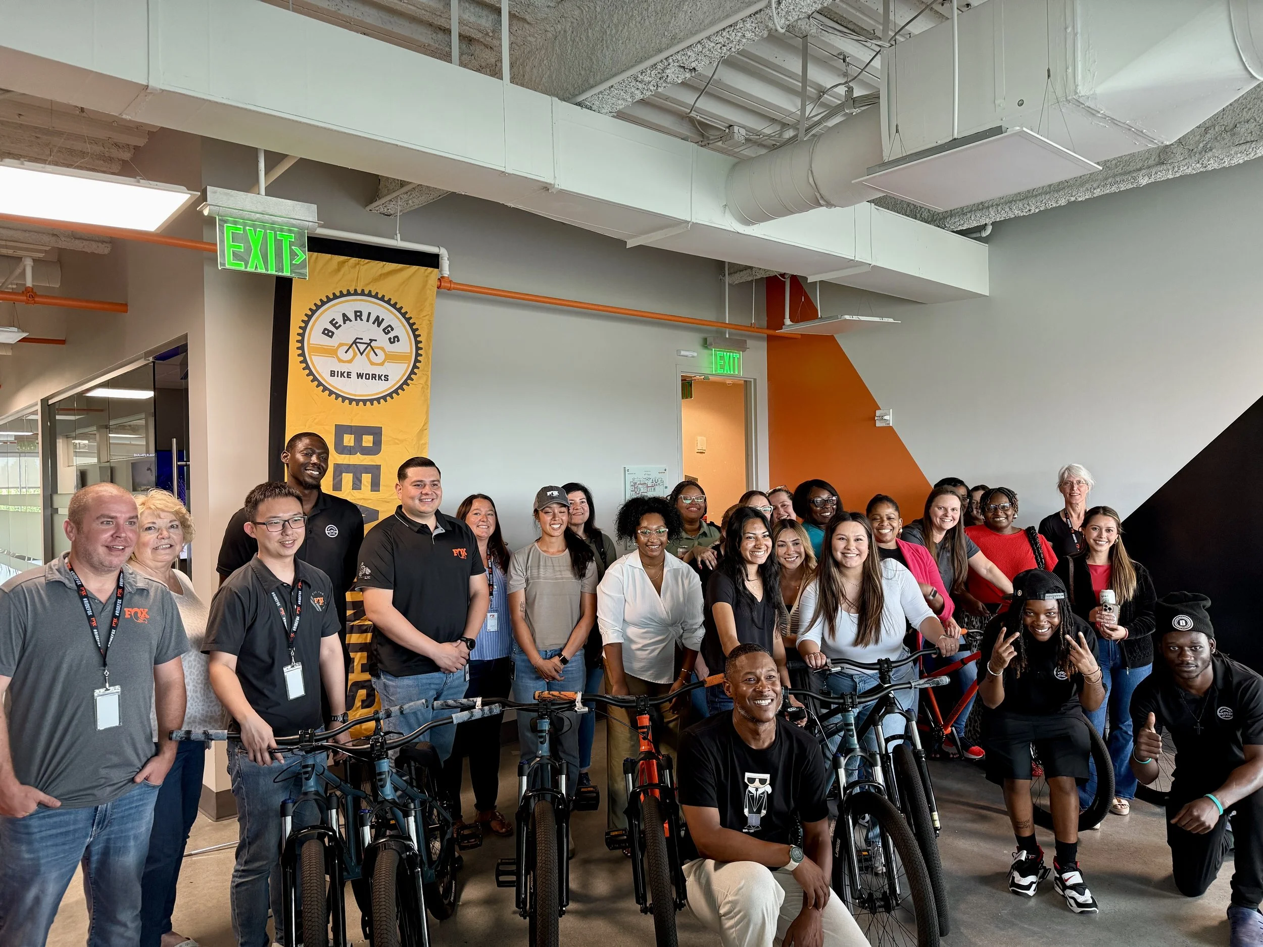 Group of diverse people gathered indoors for a photo, some with bikes, near a yellow and black banner that says 'Bearings Bike Works', in front of an exit sign.