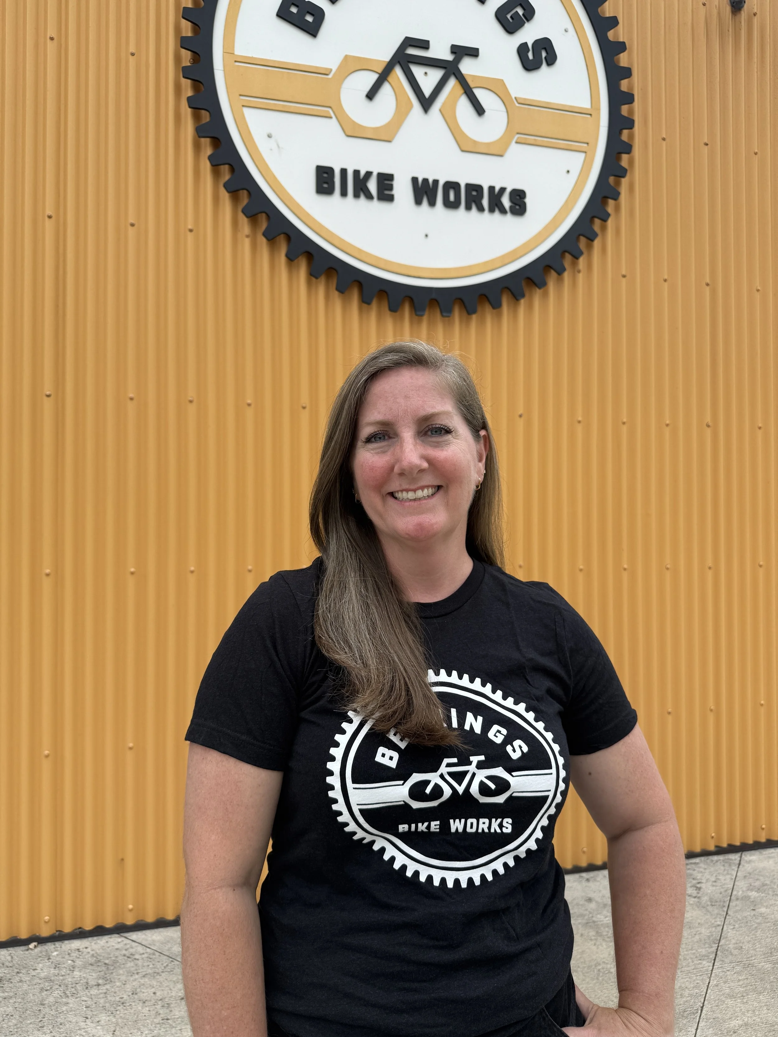 A woman with long brown hair smiling, wearing a black t-shirt with a bike-themed logo, standing in front of a yellow corrugated metal wall with a large sign that reads 'Bearings Bike Works' and a bicycle graphic.