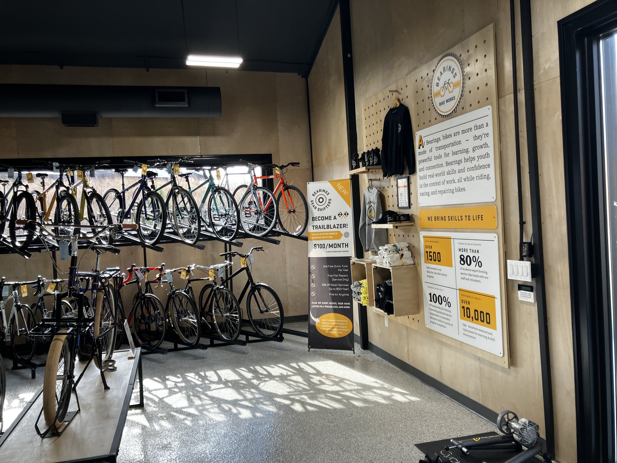 An indoor bike shop with bicycles on display, including bikes mounted on a wall rack and others on the floor. There is a wooden wall with informational posters and product shelves, with sunlight coming in through a window.