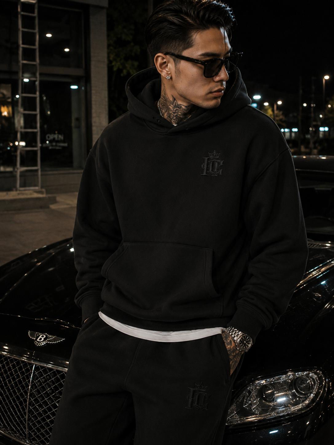 A young man wearing black sunglasses, a Inspired Culture Premium Heavyweight Hoodie with a logo, black pants with a similar logo, and a watch, stands next to a black luxury car at night.