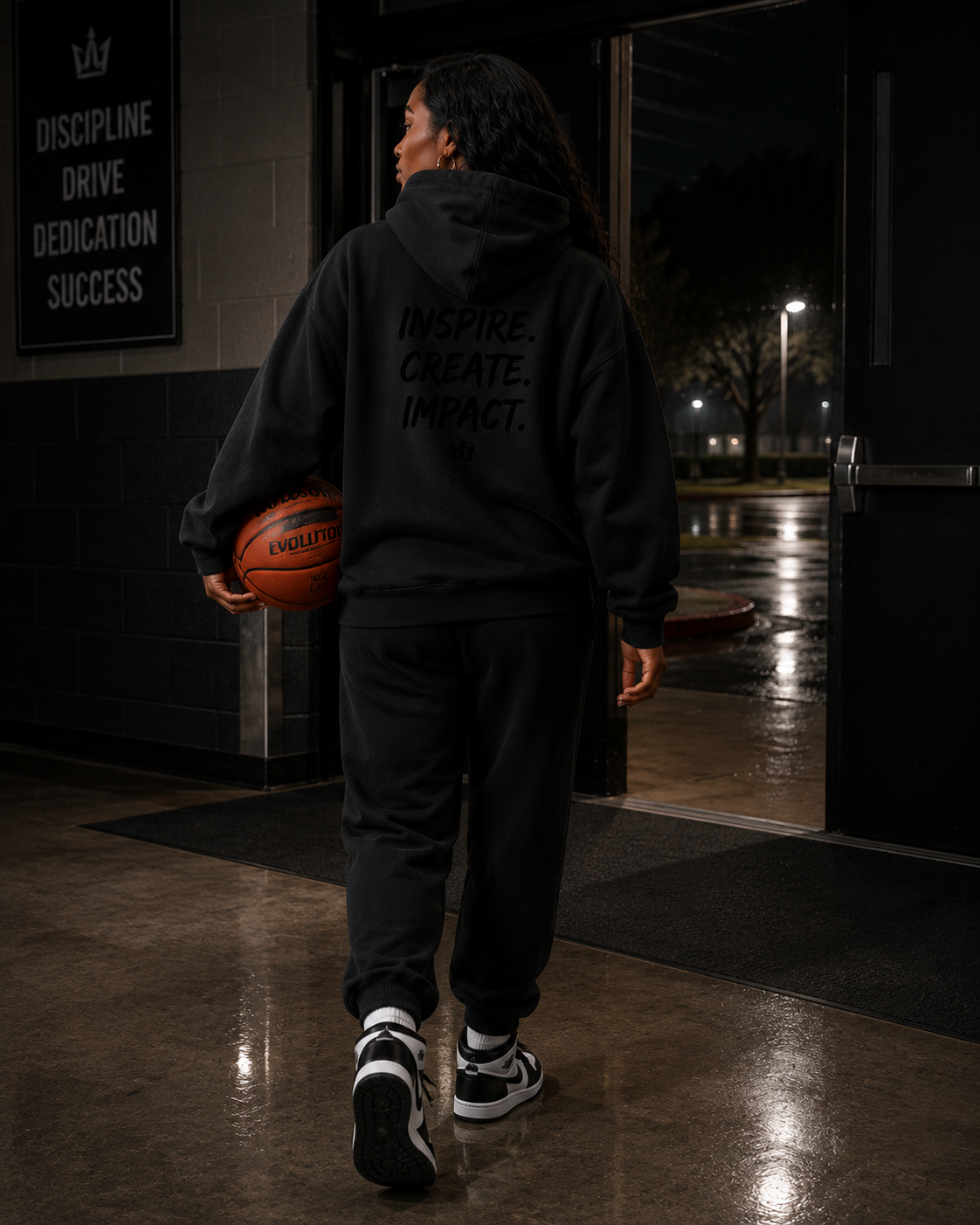 A woman wearing a black hoodie and sweatpants holding a basketball, standing at an entrance with a sign that reads, "Discipline, Drive, Dedication, Success."