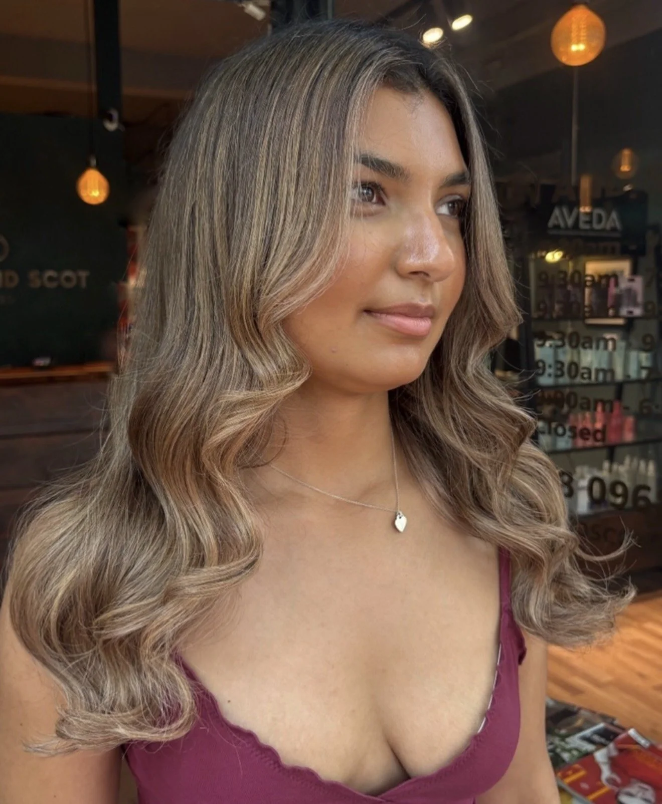 A young woman with long, wavy, light brown hair with blonde highlights, wearing a burgundy top with ruffled edges and a silver heart necklace, standing indoors with warm lighting and signs in the background.