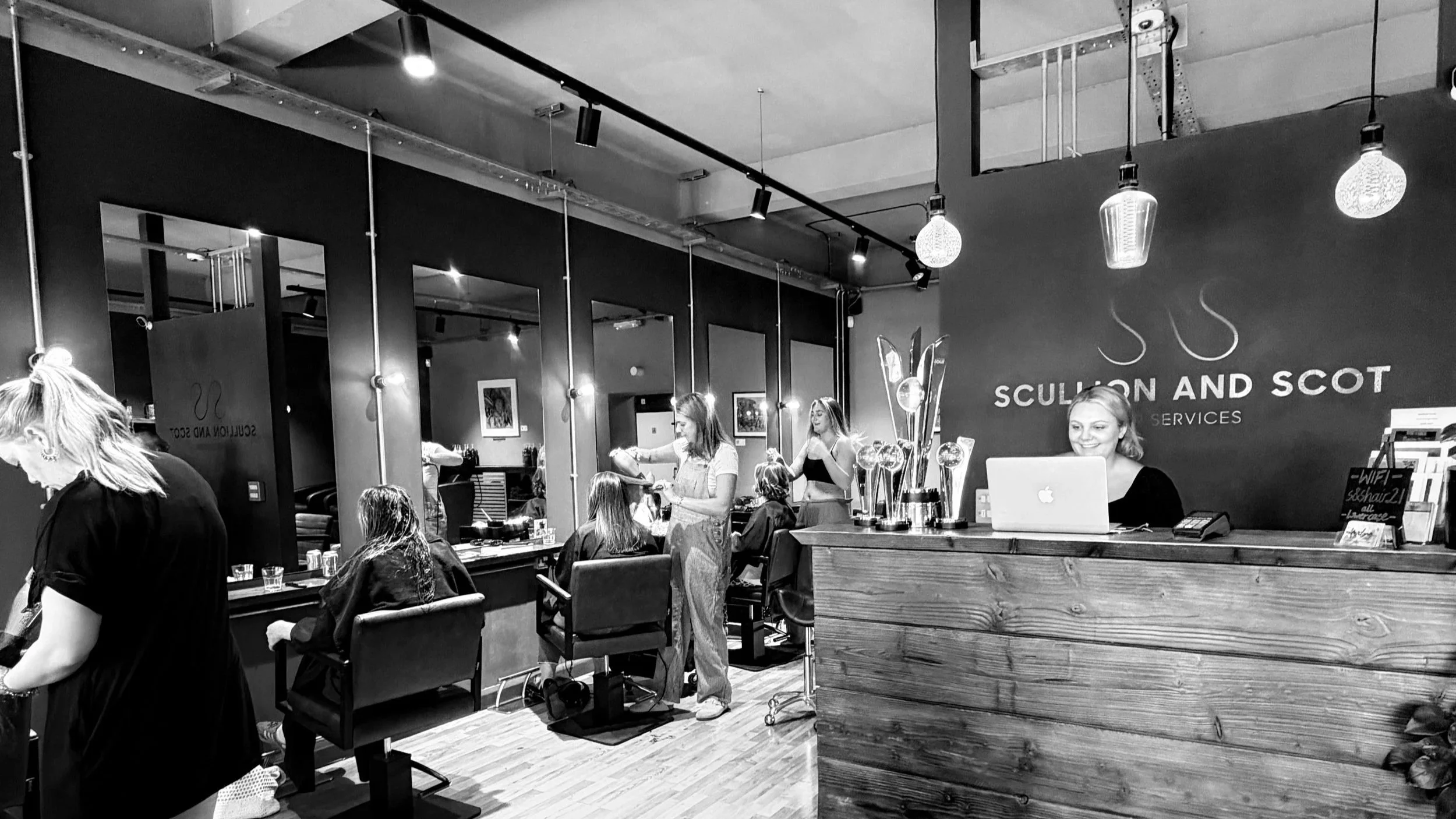 Inside a modern hair salon with clients getting hair treatments and a receptionist working on a laptop at the counter. The salon has hanging pendant lights and large mirrors.