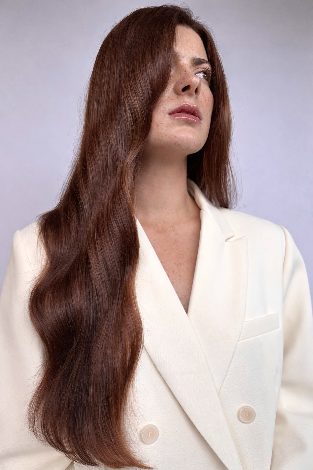 A woman with long, wavy red hair wearing a white blazer looking to her right against a plain background.