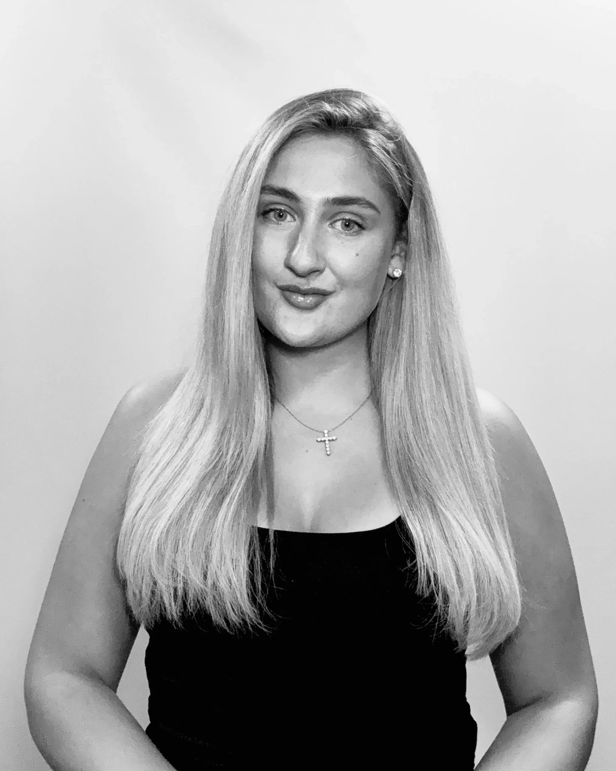 Black and white portrait of a woman with long, straight blonde hair, wearing a black top and a cross necklace, standing against a plain background.