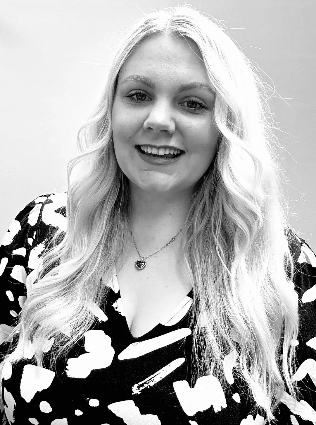 A young woman with long wavy hair, smiling, wearing a patterned top and a necklace with a heart-shaped pendant, in a black-and-white photograph.