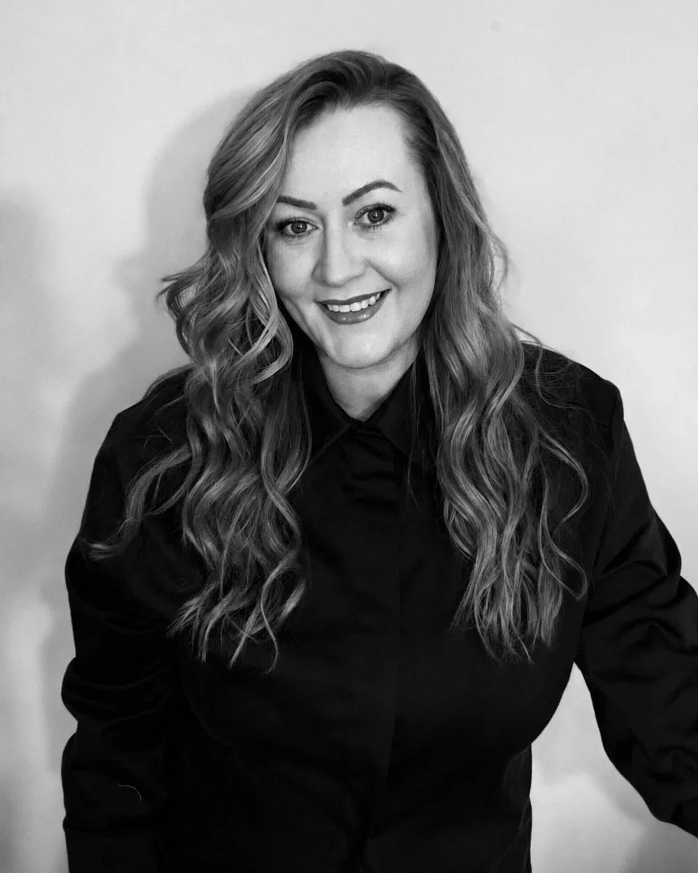Black and white photo of a woman with long wavy hair, smiling, wearing a dark shirt, posed against a plain light background.
