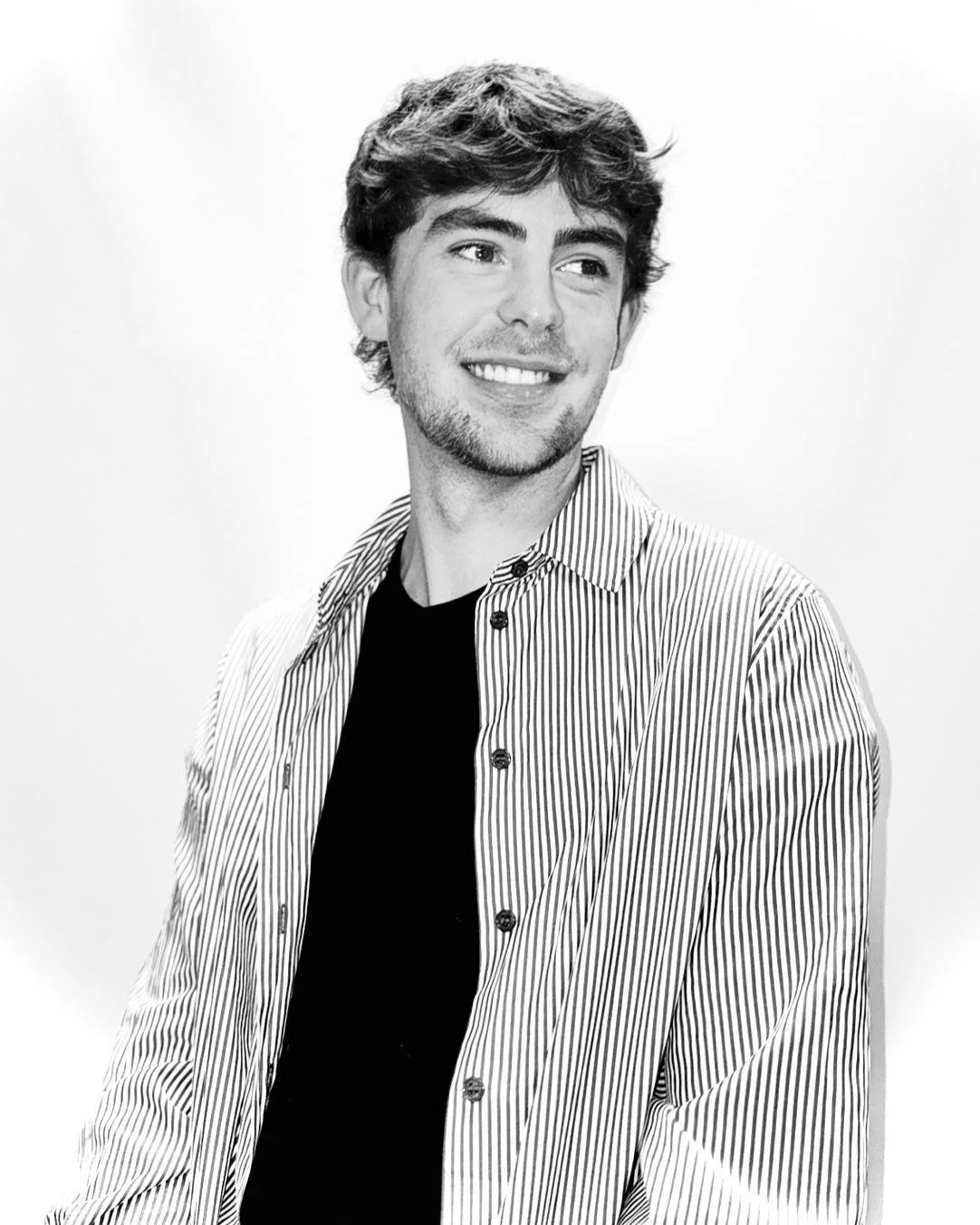 Black and white photo of a young man with wavy hair and a beard smiling, wearing a striped shirt over a black t-shirt, standing against a light background.