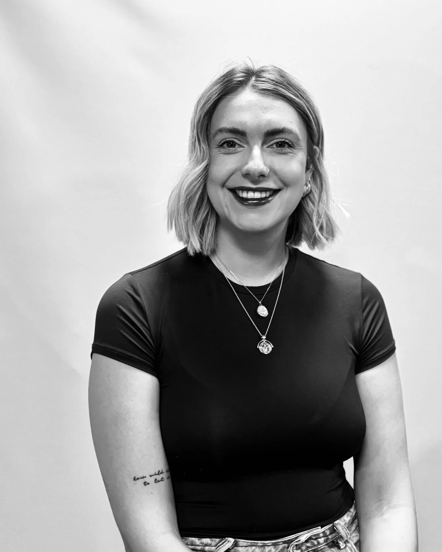 Black and white portrait of a smiling woman with shoulder-length hair, wearing a black top, layered necklaces, and earrings, standing against a plain light-colored background.