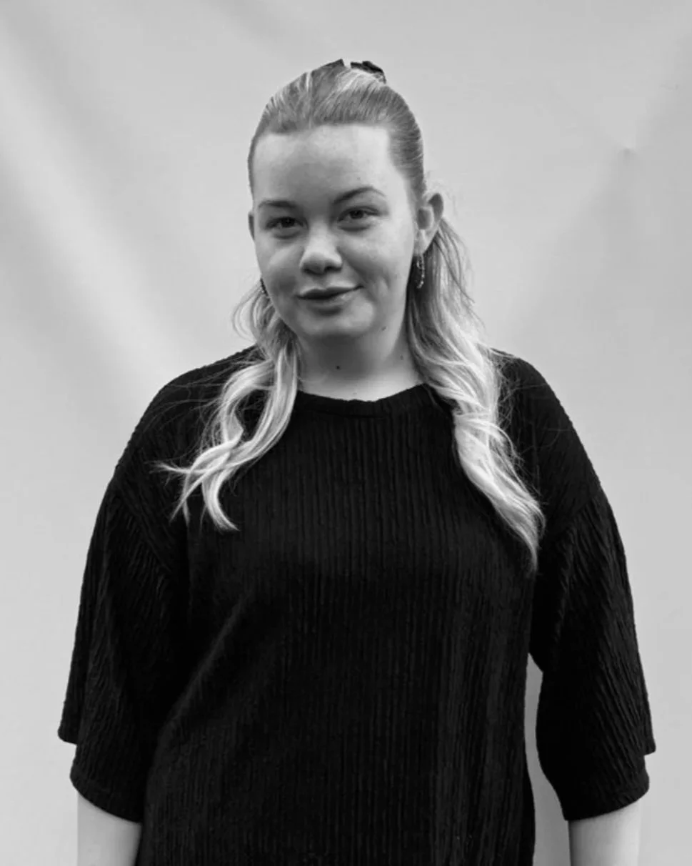 Black and white photo of a young woman with wavy, shoulder-length hair, wearing a dark, long-sleeved top, standing against a plain light background.