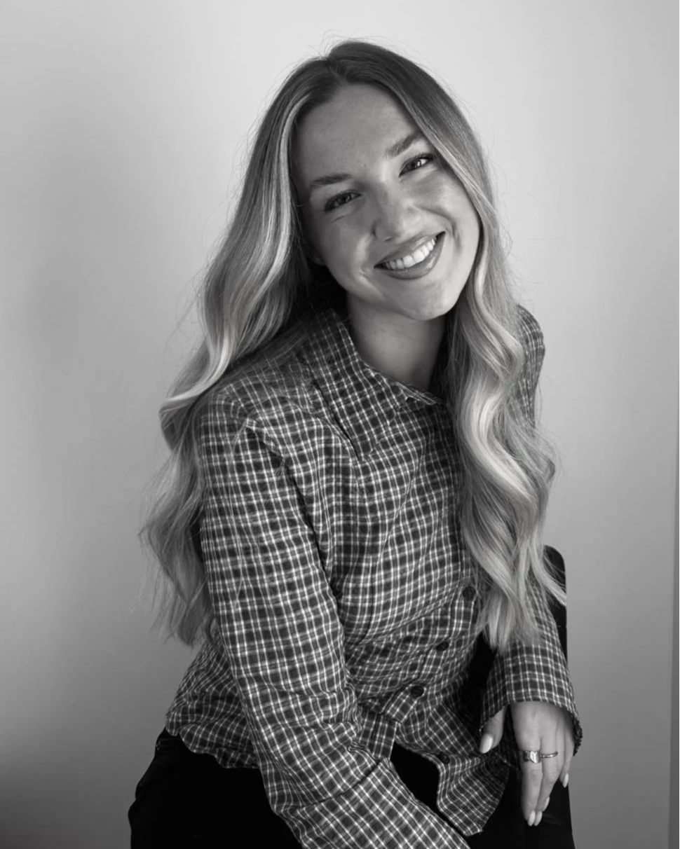Black and white portrait of a young woman with long wavy hair, smiling and wearing a checkered shirt, seated with her hands resting on her lap against a plain background.