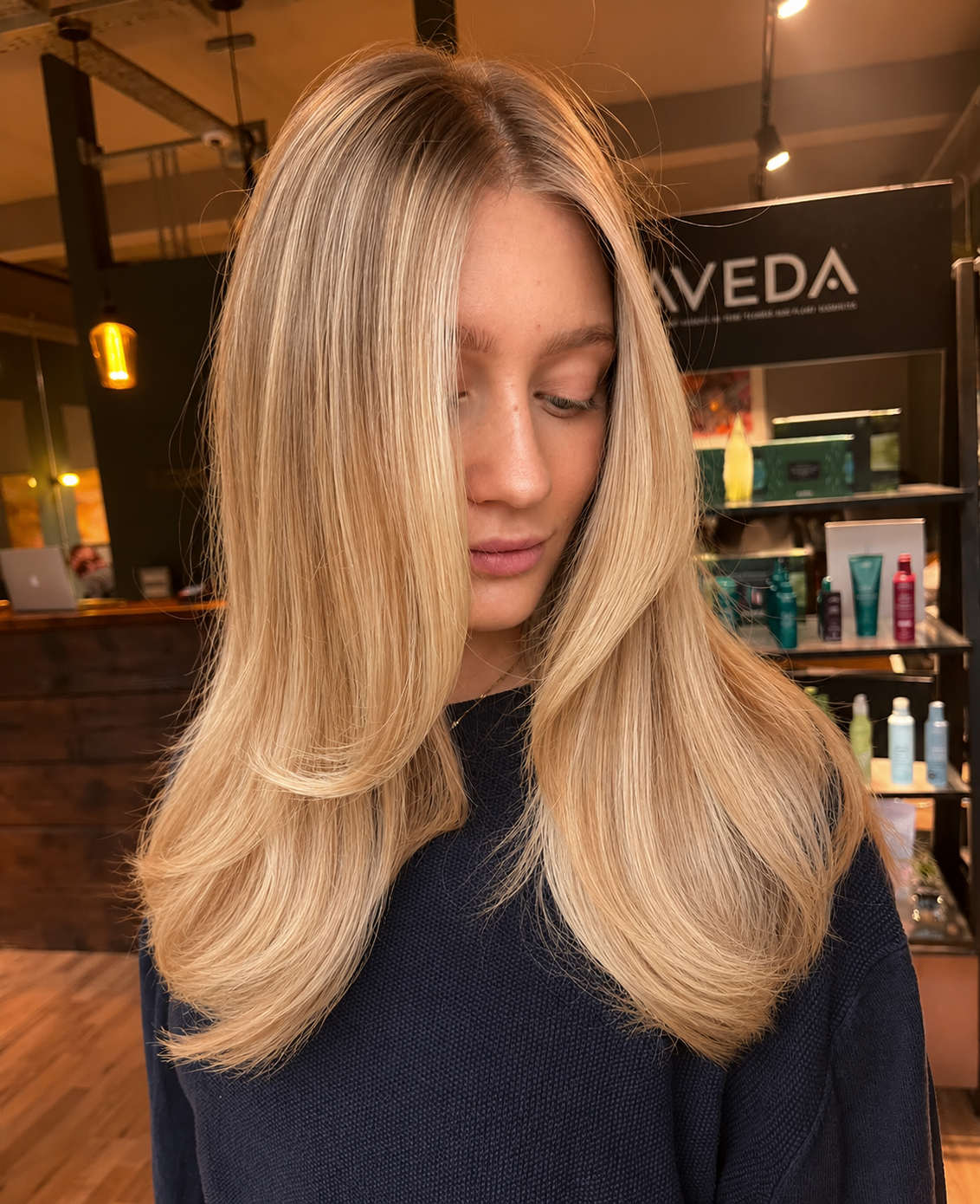 A woman with long, straight blonde hair, wearing a dark top, standing in a modern salon or retail store with shelves of hair and skin care products in the background.