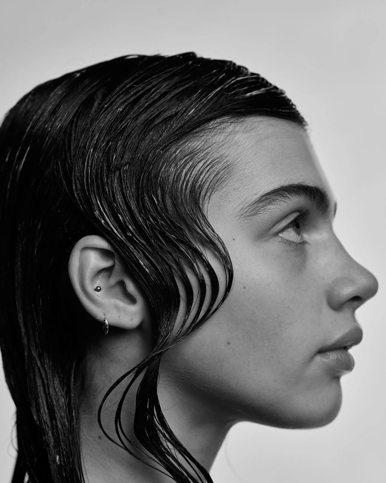Black and white close-up side portrait of a young woman with wet, slicked-back hair, ear piercings, and a calm expression.