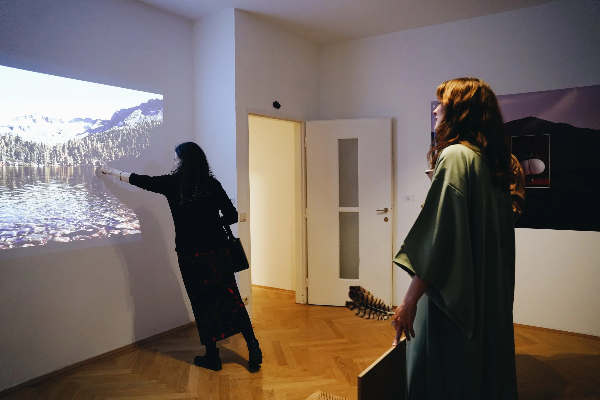 Two women in an art gallery, one pointing at a large projected image of a mountain lake scene, the other observing. The room has wooden flooring and white walls, with a door in the background.