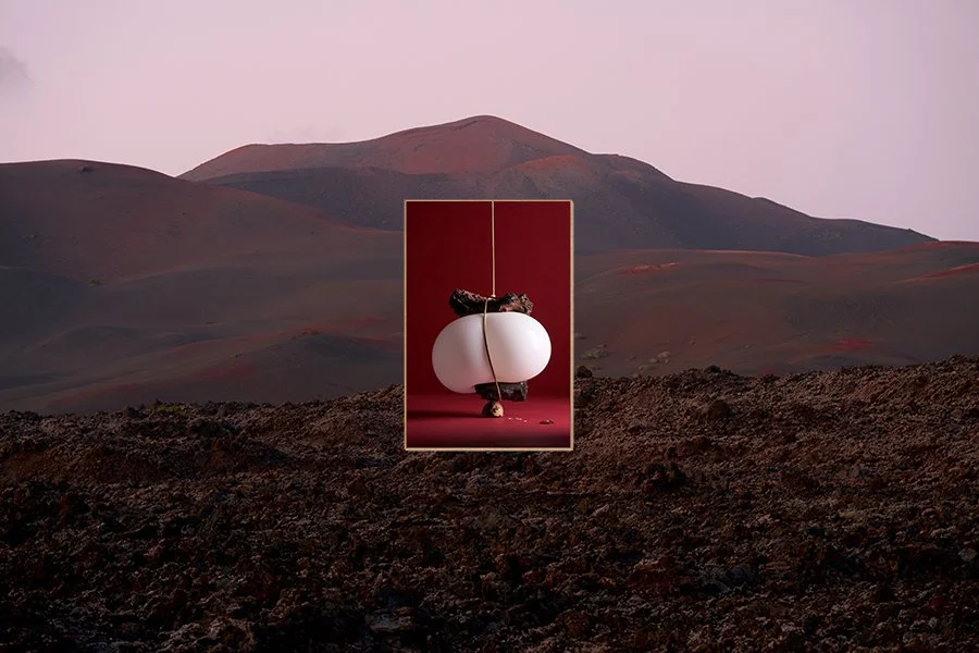 A framed photograph of a white, modern art sculpture resembling a pair of rounded shapes, set against a red background, with rocks and rolling hills in the distant landscape.