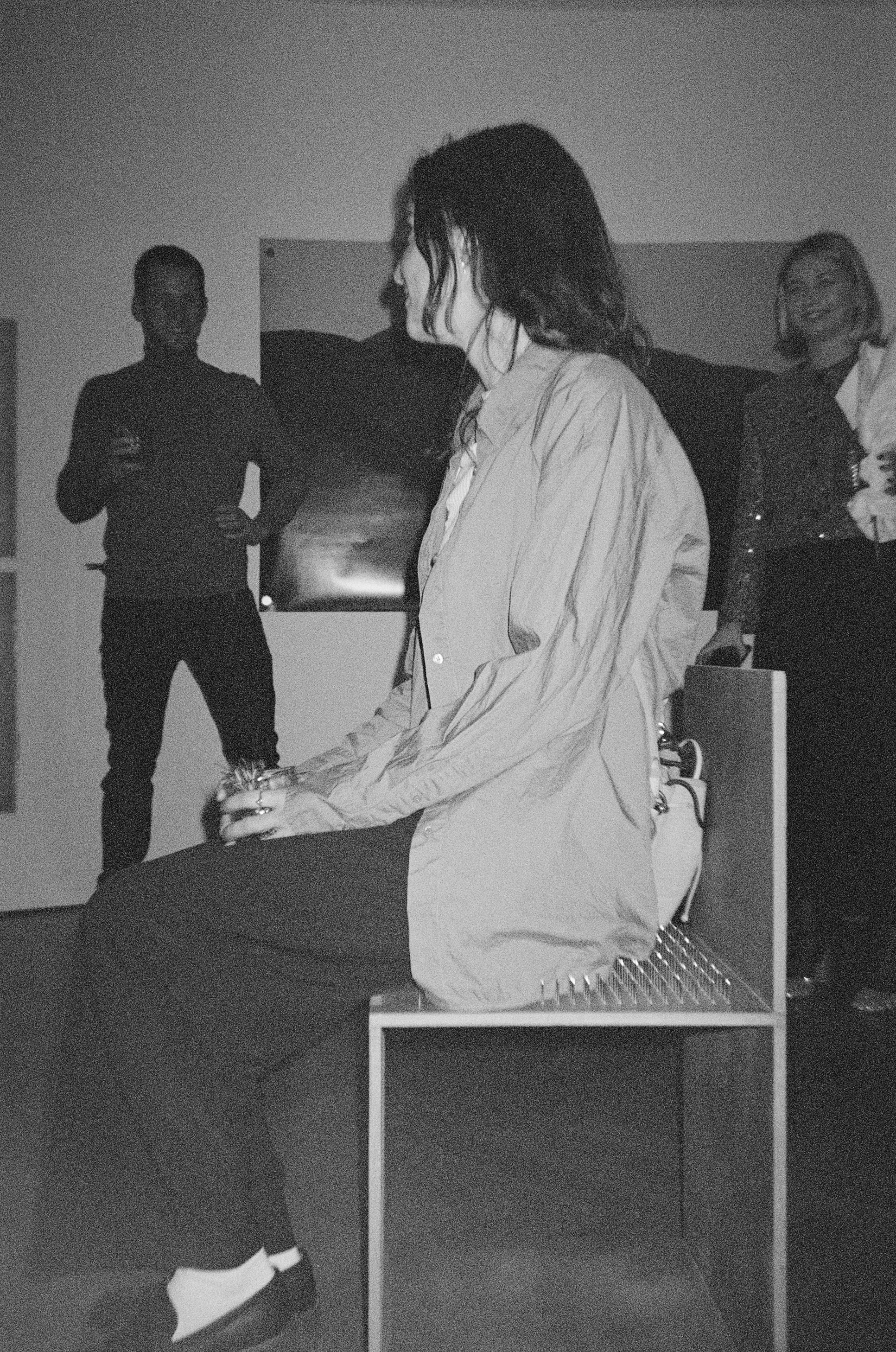 Woman sitting on a bench with two women and one man standing behind her in a room with a wall art, all in black and white.