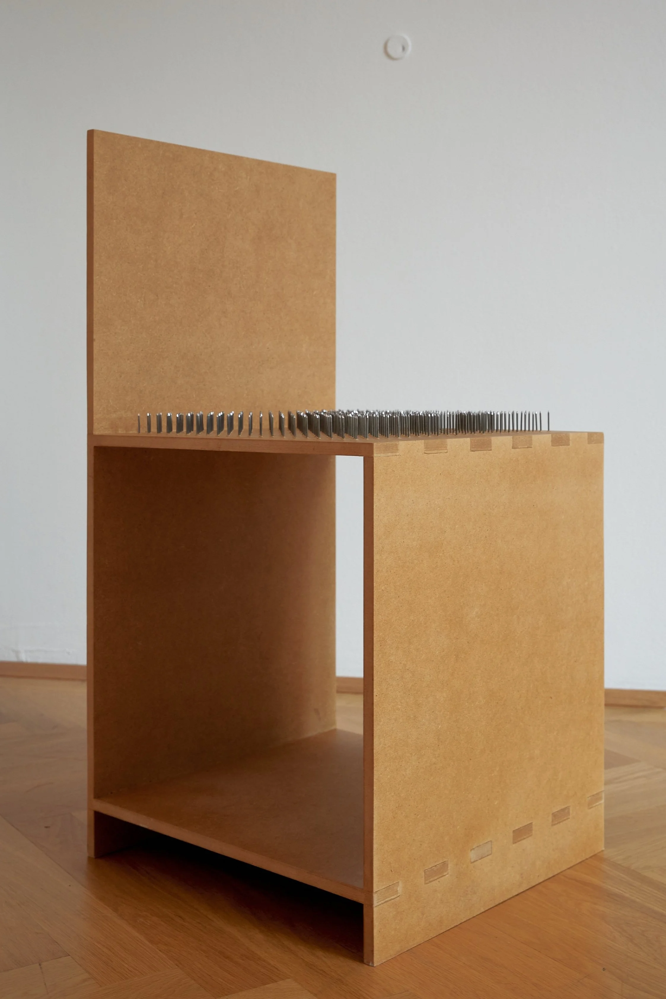 A wooden display case with a back panel, featuring rows of metal nails on top, placed on a hardwood floor, with a plain white wall in the background.