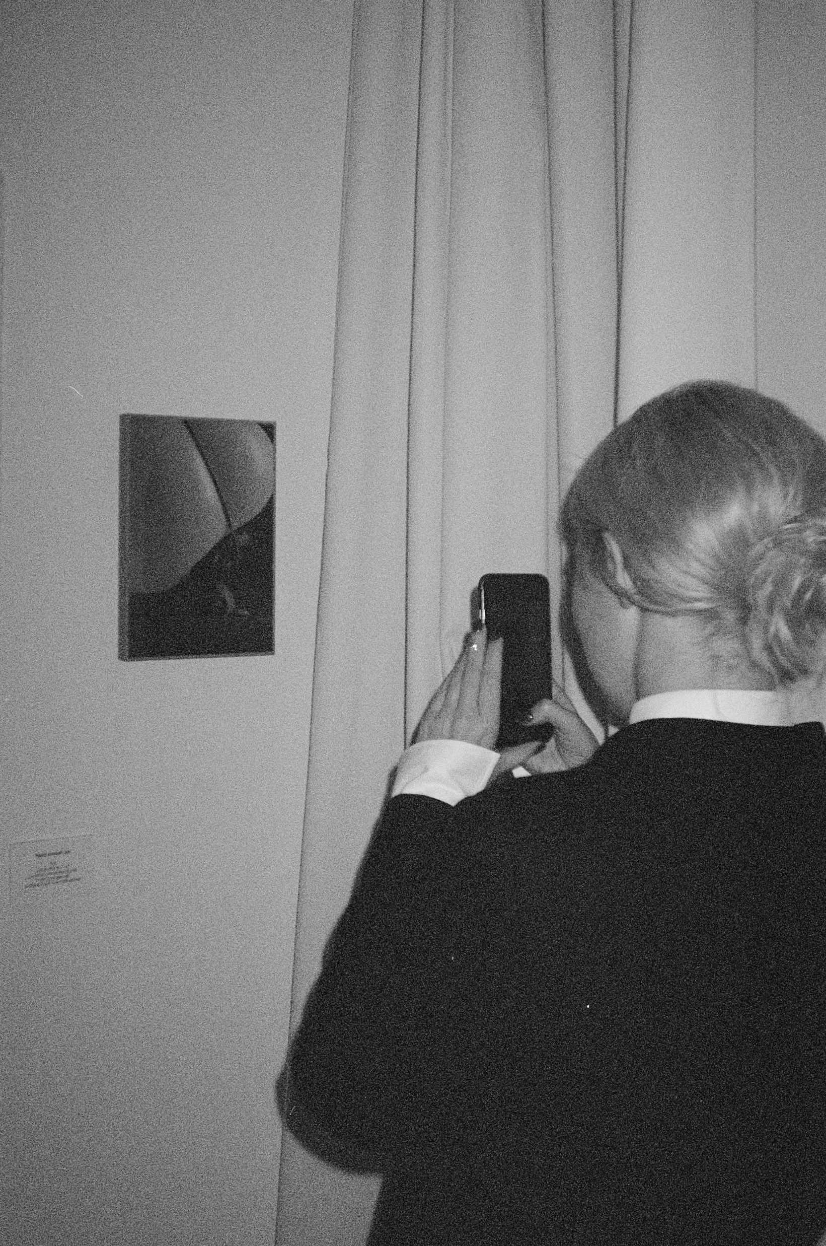 A woman with styled hair taking a photo of an artwork on a wall in a gallery or museum, black and white photo.