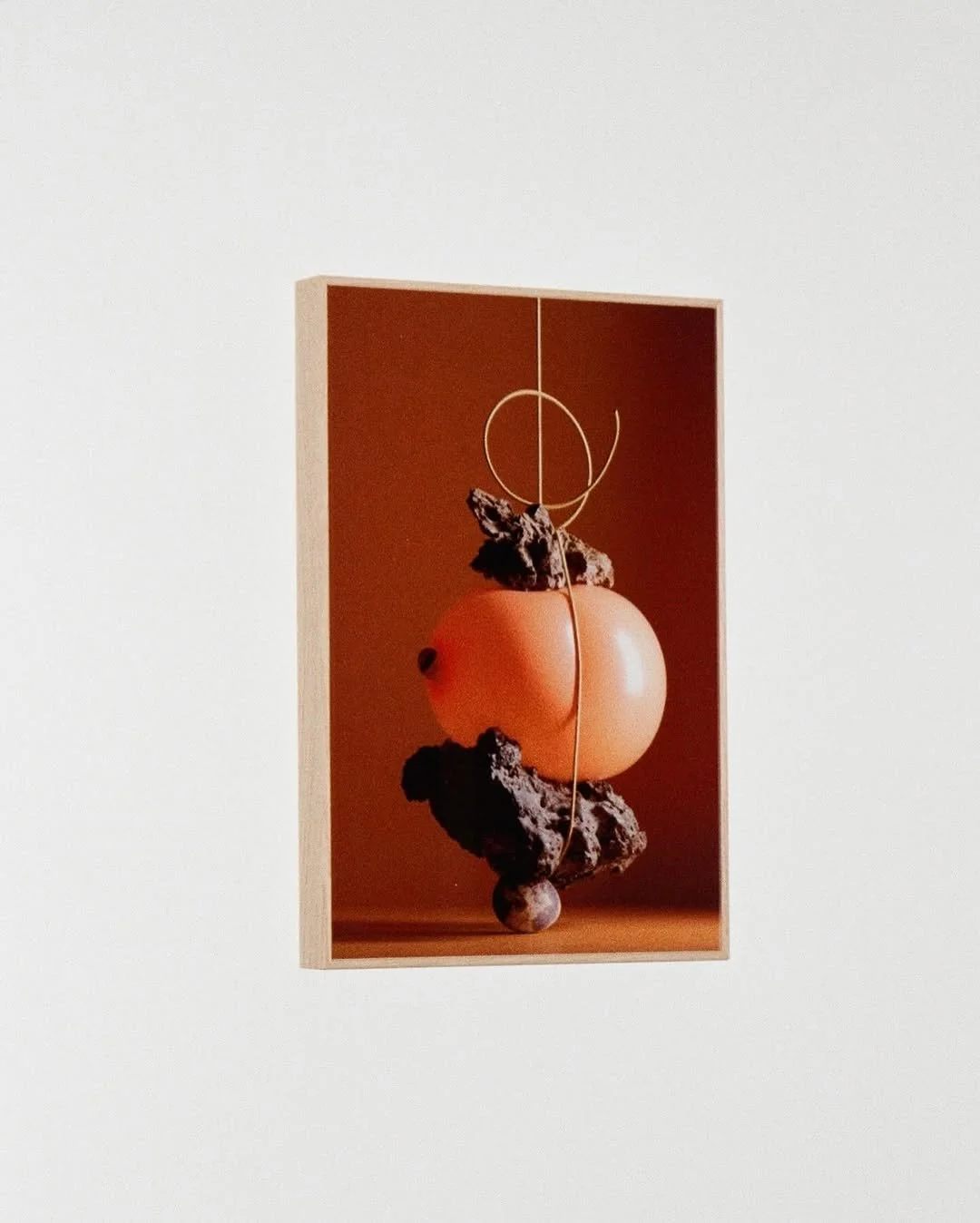 Abstract art featuring a large peach-like structure balanced on dark, irregular rocks with a small gray sphere at the bottom. Thin metal wire loops above the peach, set against a brown background framed with a light wood border.