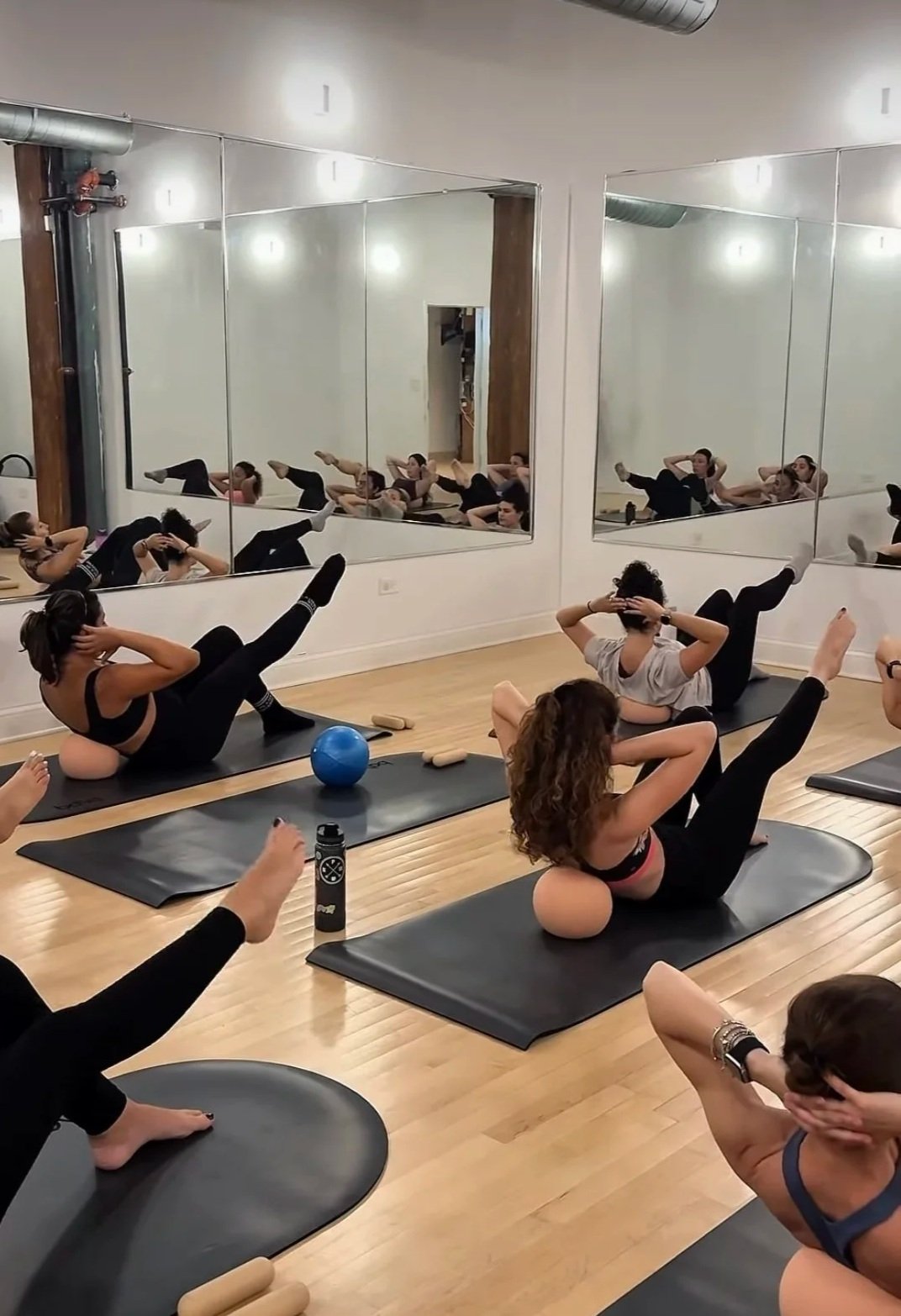 Women participating in a fitness class in a studio with large mirrors, exercising on mats with foam rollers, a blue exercise ball, and water bottles visible.