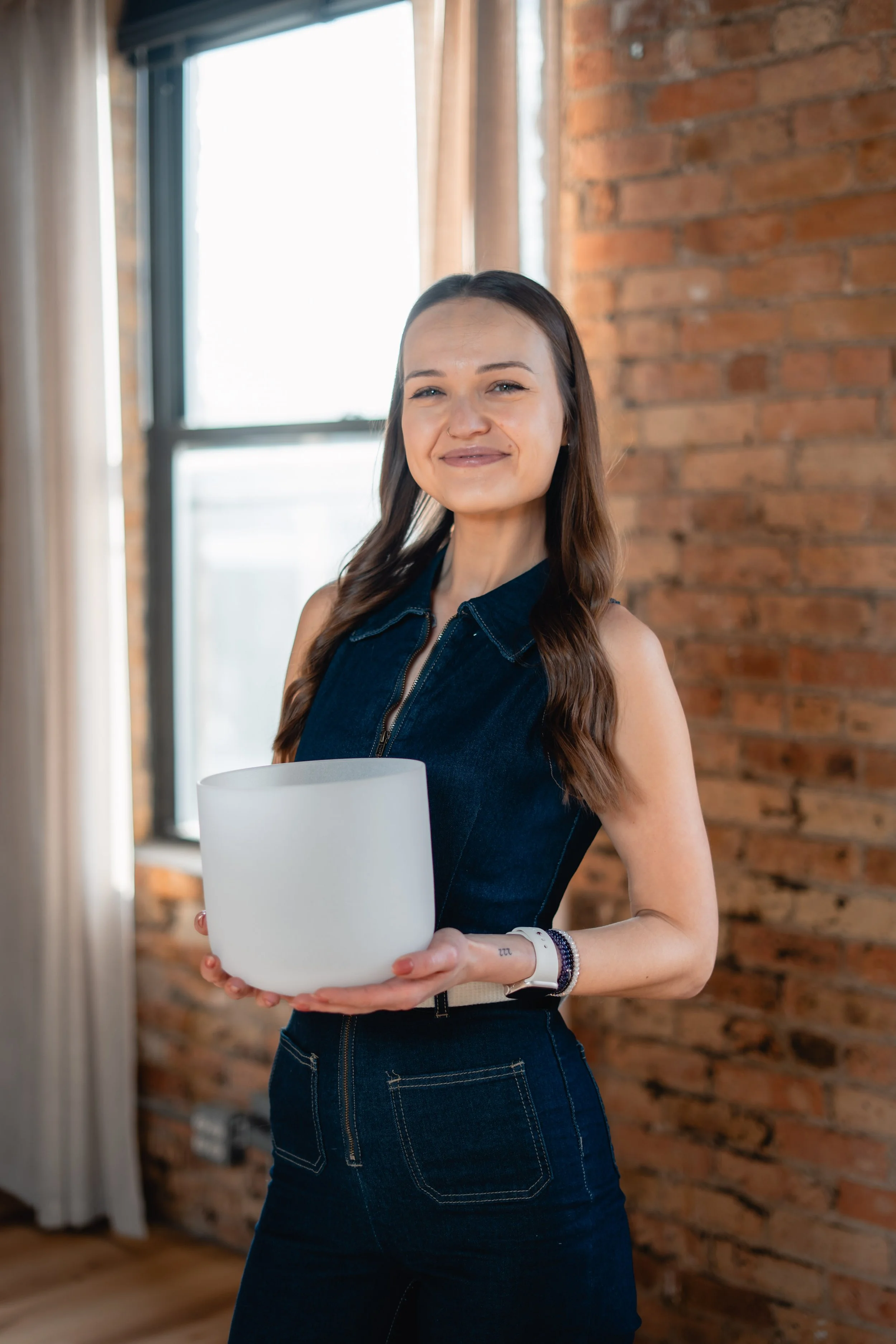 A woman with long brown hair and a denim sleeveless top standing indoors, holding a large white candle, with a brick wall and large window in the background.