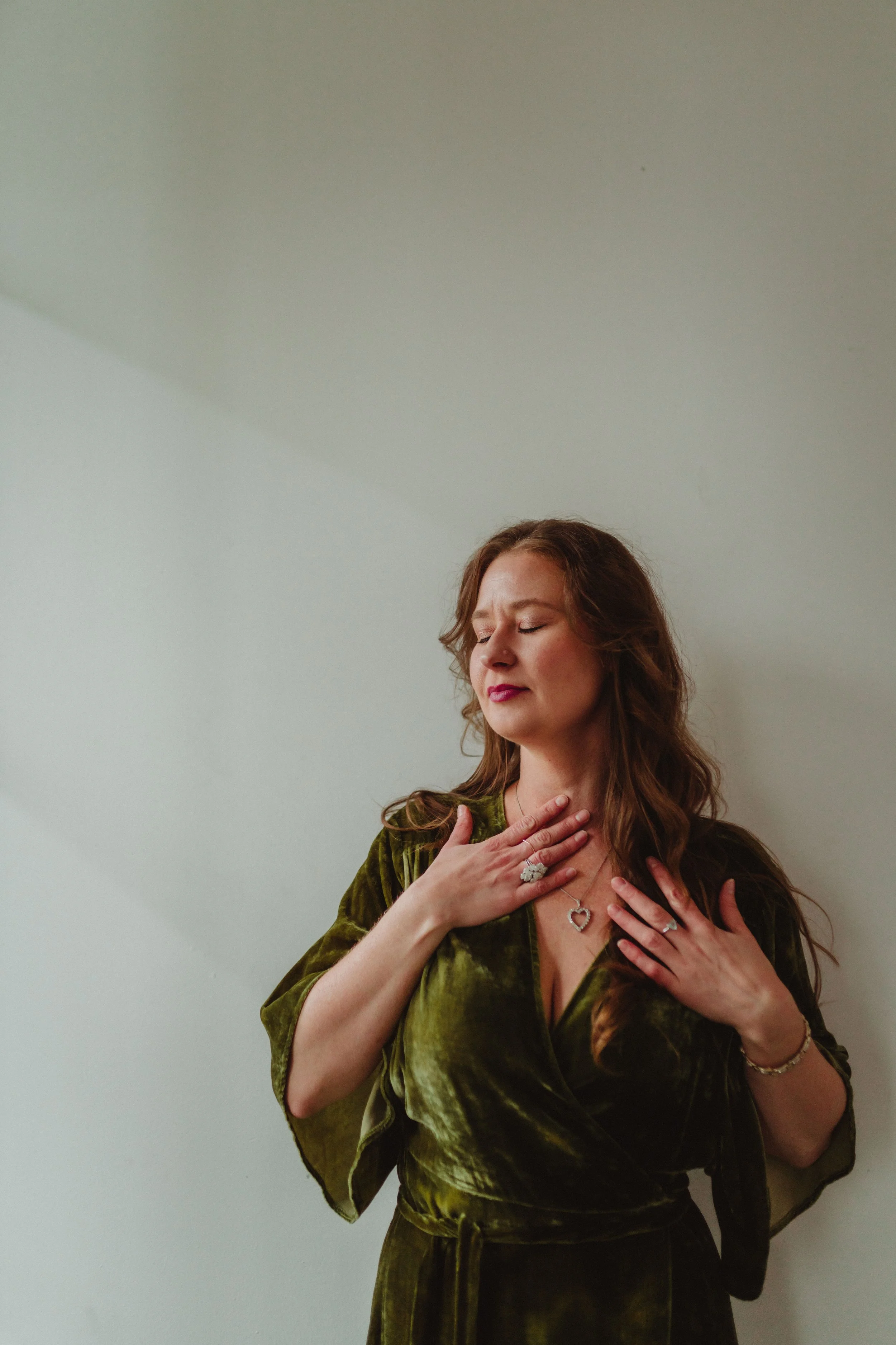 Woman with long brown hair wearing an olive green dress, jewelry, and rings, standing against a white wall, with her eyes closed, touching her chest.