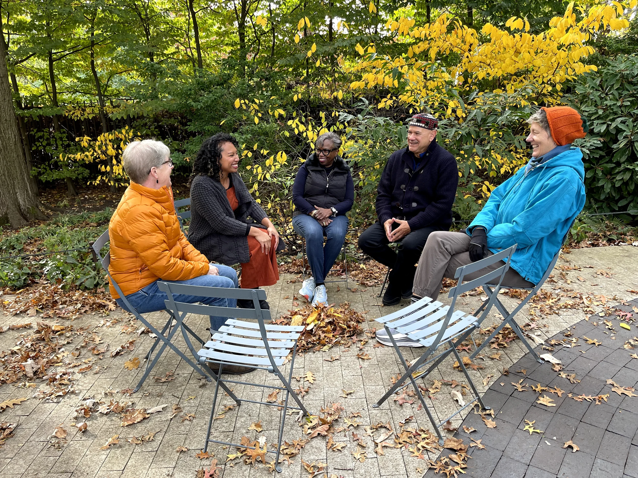 Five people sitting in a circle outdoors on a paved area, engaged in conversation. Four are women and one is a man. They are dressed warmly, wearing jackets, scarves, and a beanie, indicating cool weather. The background features trees with green and yellow foliage, and fallen leaves on the ground, suggesting autumn.