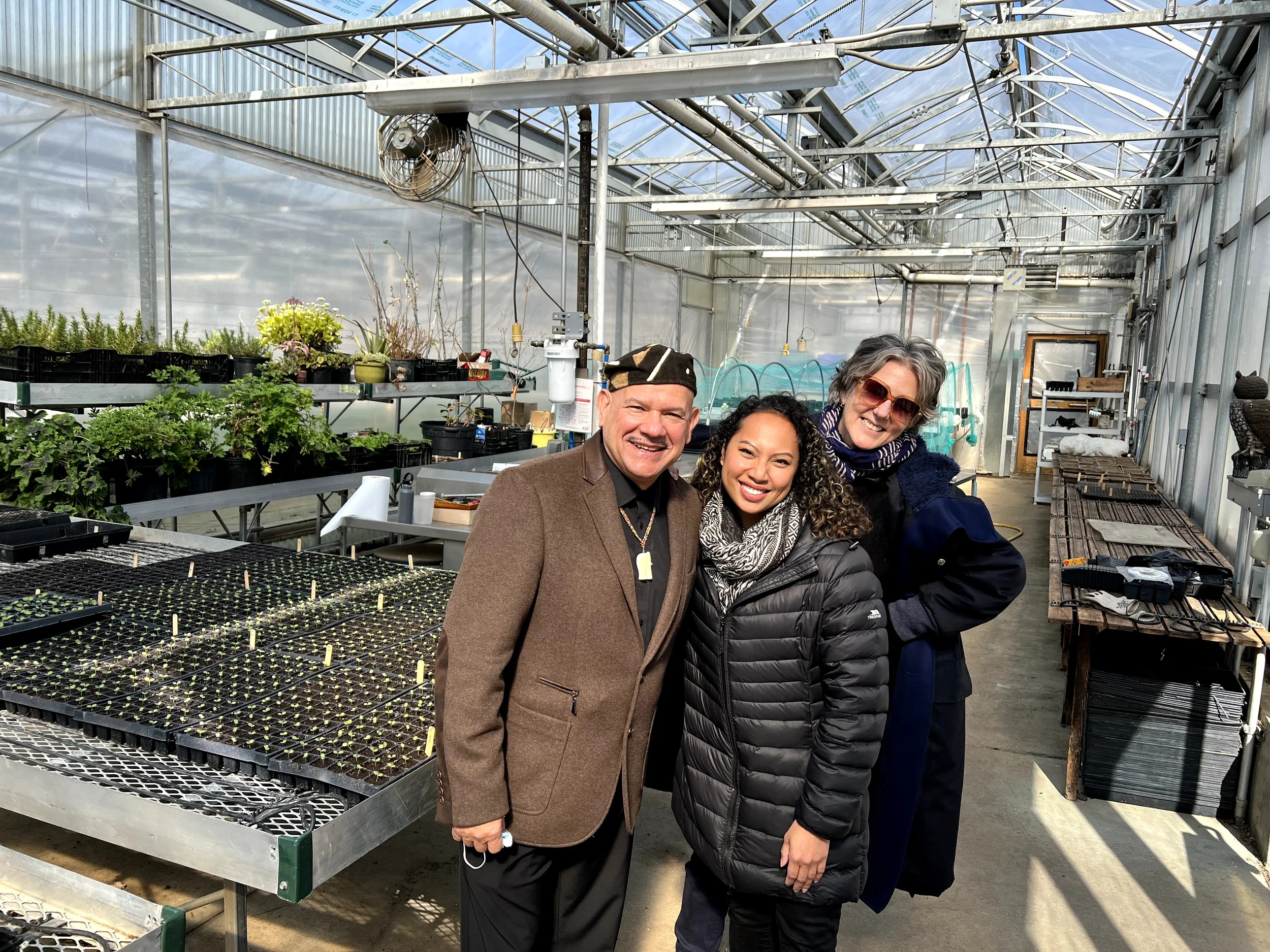 Three smiling people standing inside a greenhouse with plants on shelves and trays of seedlings on tables.