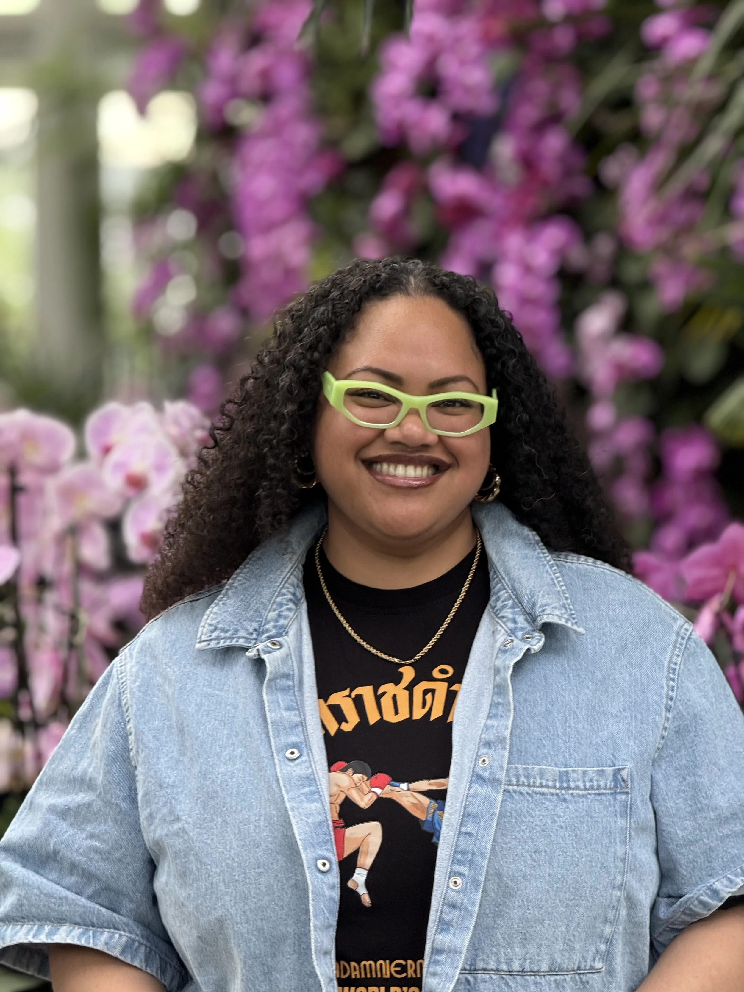 A woman with curly dark hair green eyeglasses, denim shirt in front of orchids.