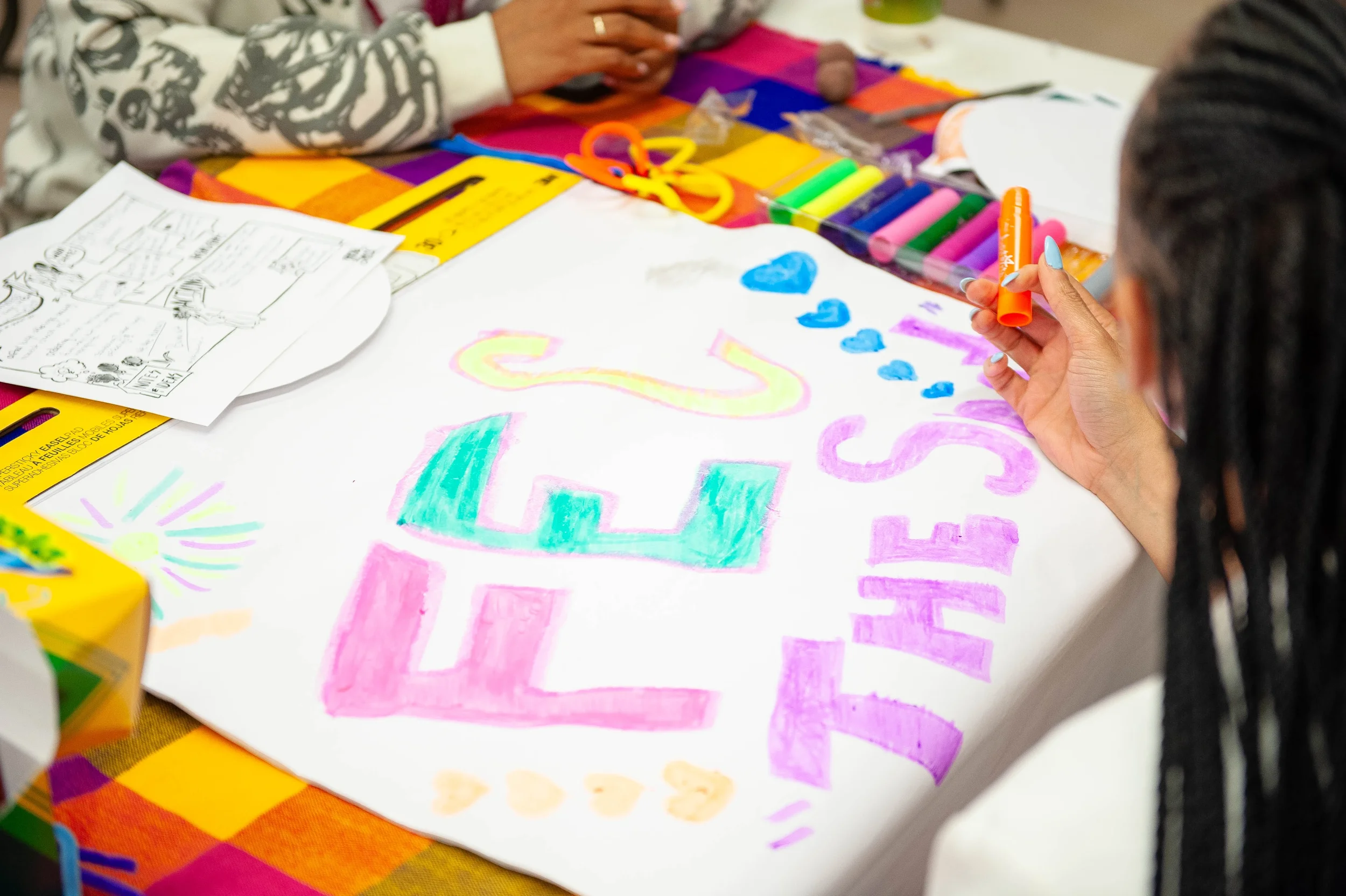 Person decorating a colorful poster with markers, words, and hearts on a table with art supplies.