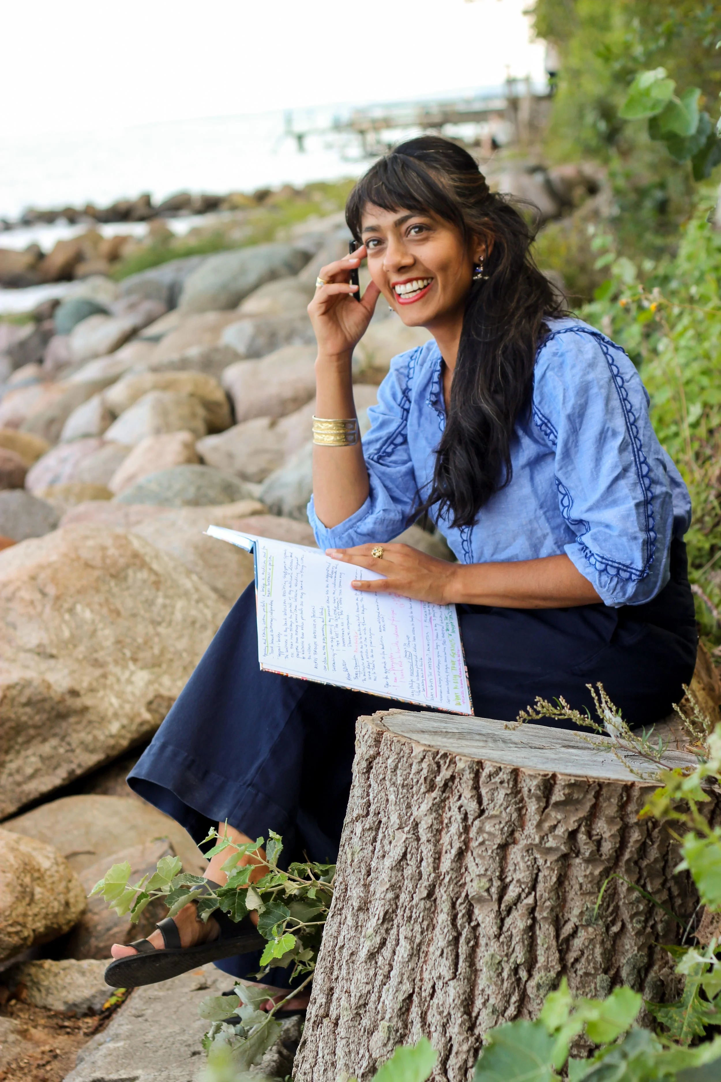 A woman sitting on a tree stump near a rocky shoreline, talking on a cell phone, smiling, with a notebook in her lap and green leaves around her.