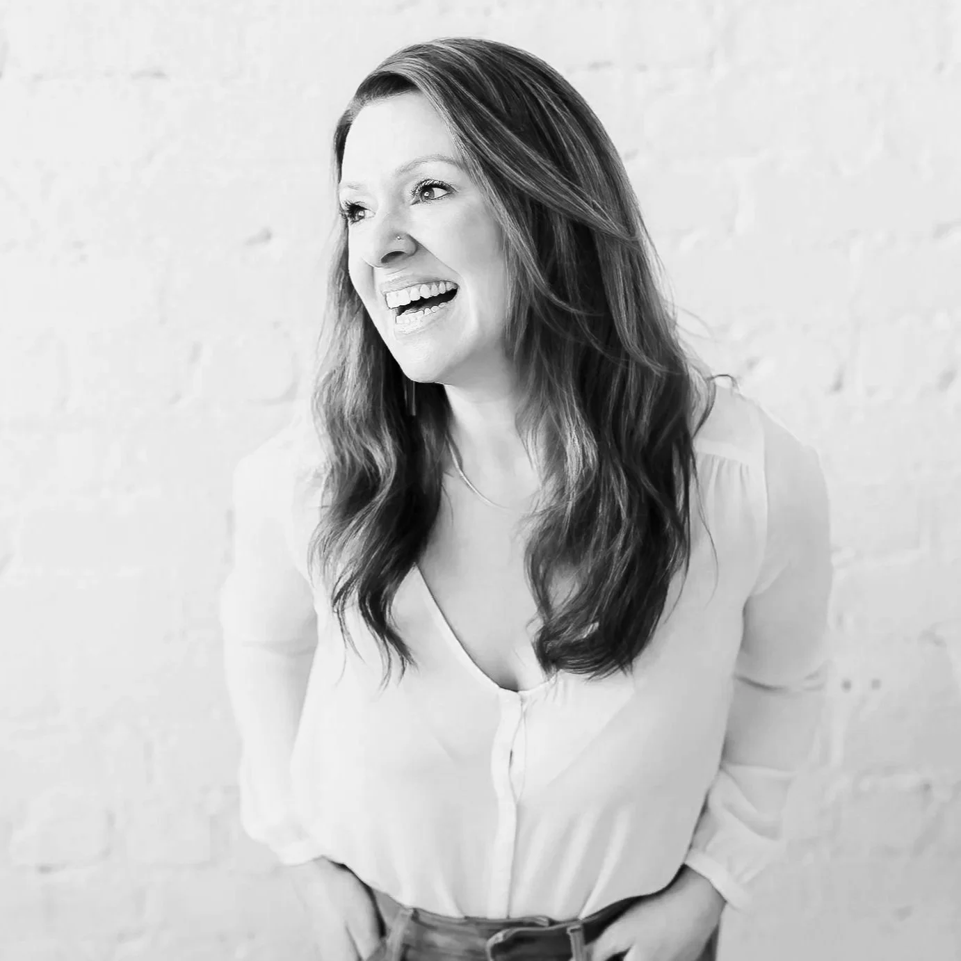 Black and white photo of a young woman smiling, sitting on a wooden chair against a white brick wall. She has long, wavy hair, large hoop earrings, and is wearing a black top and ripped jeans.