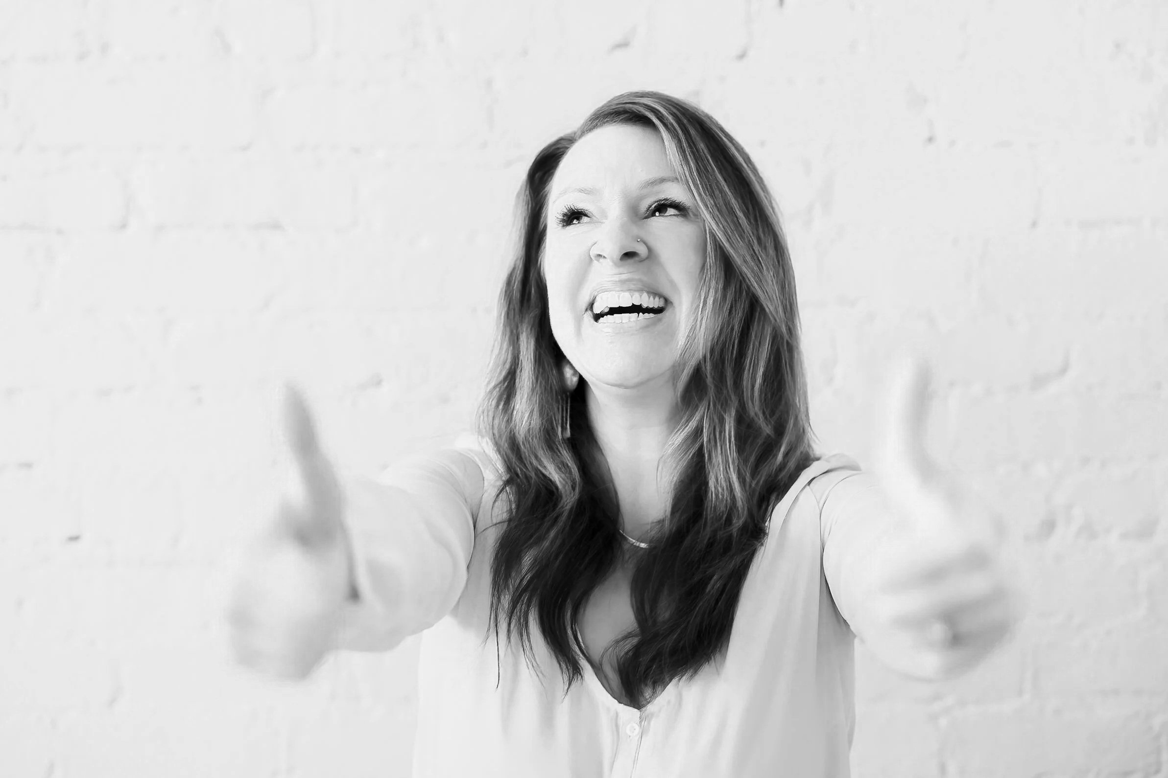 A woman with shoulder-length hair laughing, wearing a black top and light-colored jeans, touching her head with one hand, standing behind a chair against a white brick wall.