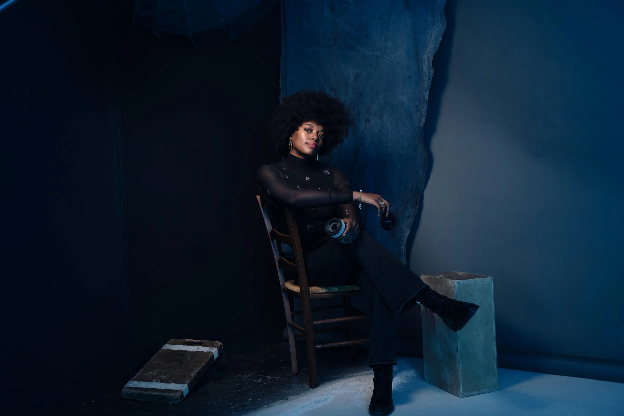 A woman with glasses and short curly hair smiling while sitting on a brown leather sofa, with a large plant behind her.