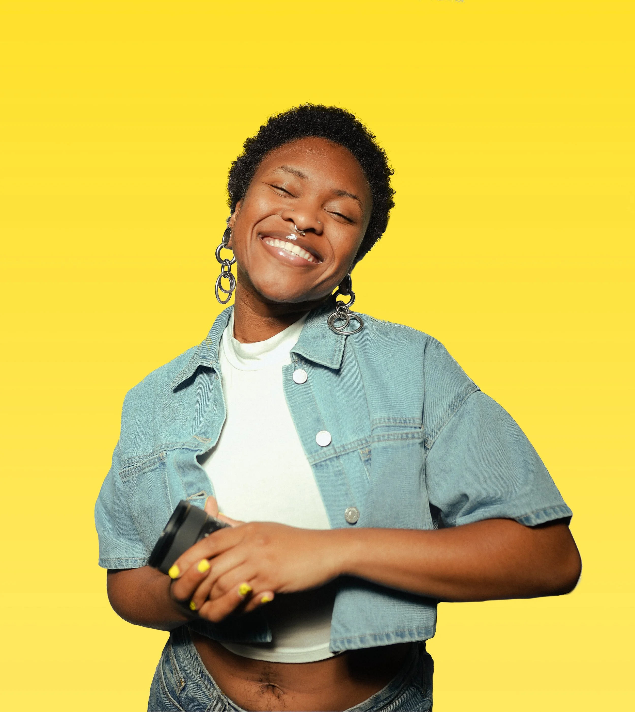 A young woman with short curly hair smiling happily, holding a phone in her hands, wearing a denim jacket and earrings, against a yellow background.