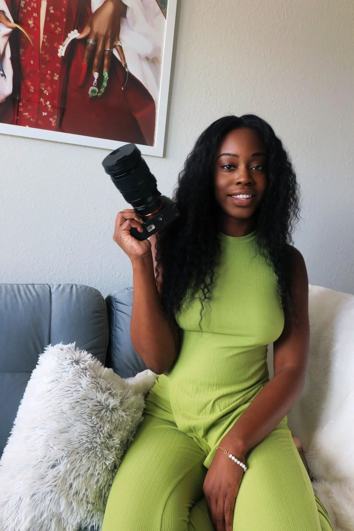A woman with long curly black hair sitting on a light-colored couch, holding a camera with a long zoom lens, wearing a bright green sleeveless outfit, smiling, with fluffy pillows behind her and a framed photo of a woman with colorful nails on the wall.