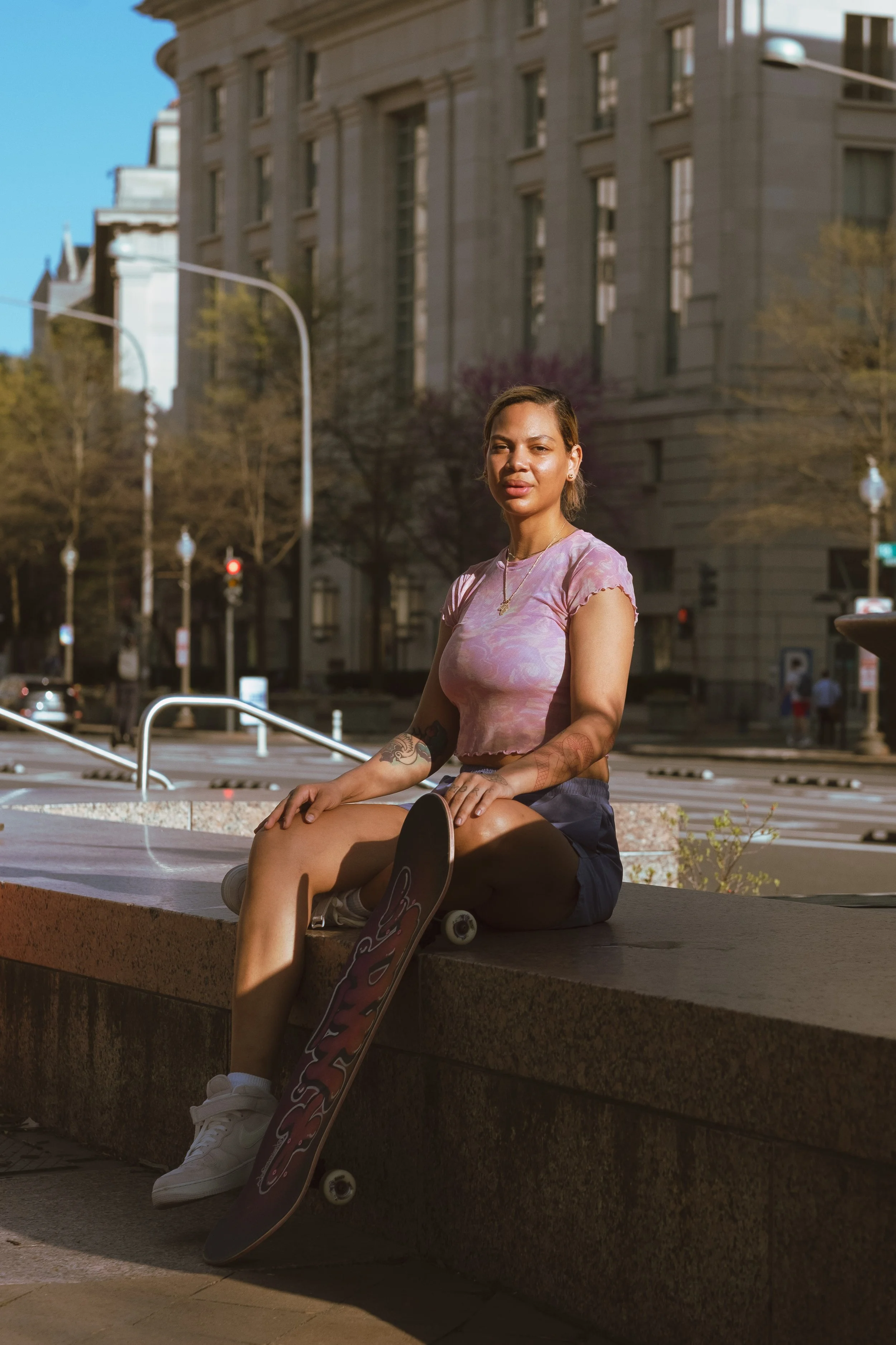 A woman sitting on a low wall holding a skateboard, with an urban city street and large buildings in the background.