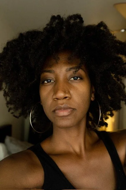 Close-up of a woman with natural curly hair, wearing large hoop earrings and a black tank top, looking directly at the camera.