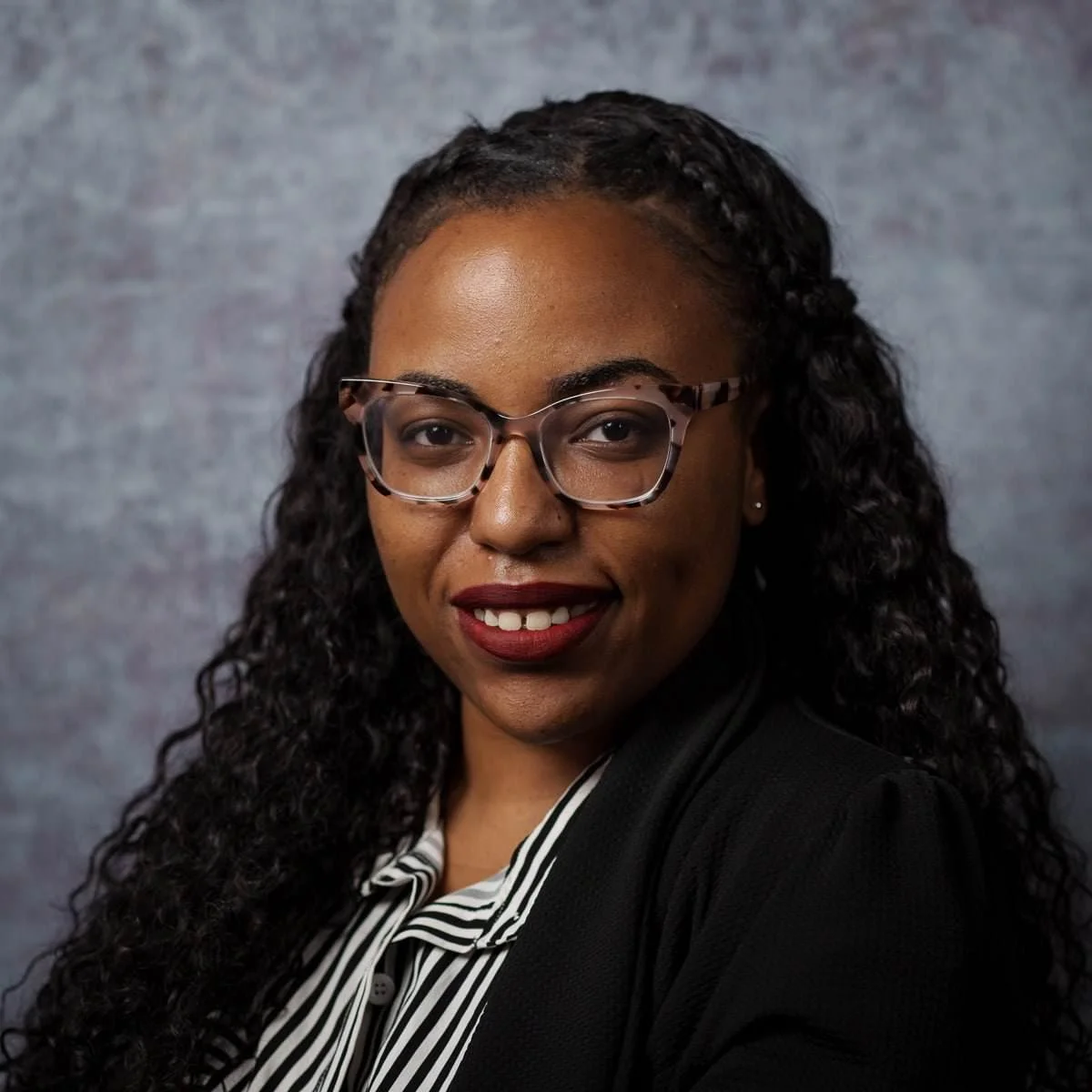Close-up of a woman with natural curly hair, wearing large hoop earrings and a black tank top, looking directly at the camera.